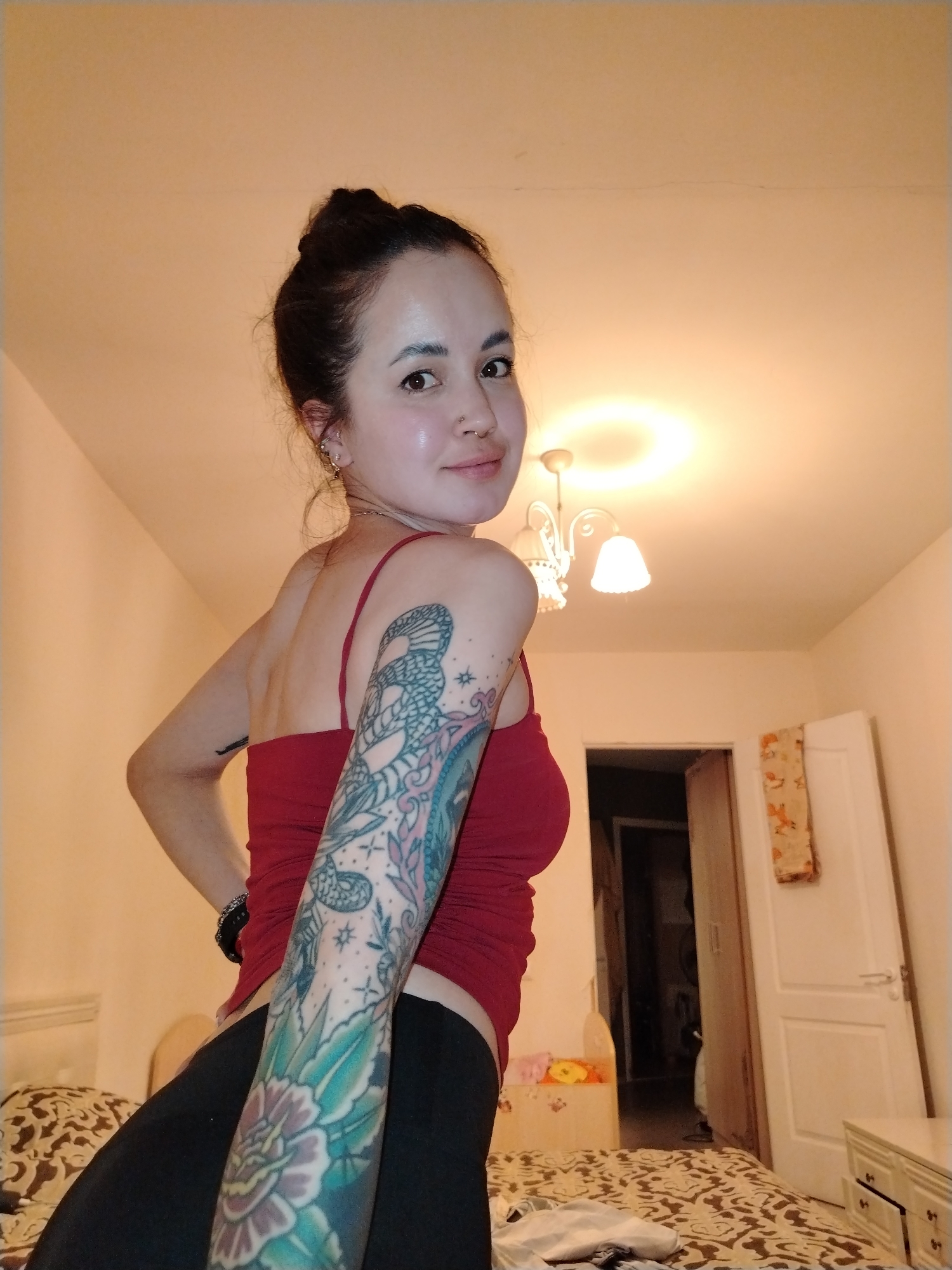 A woman with dark hair pulled into a bun is indoors in what appears to be a bedroom. She is wearing a red tank top and black pants, and has extensive tattoos on her left arm. She is looking over her shoulder at the camera with a slight smile. A bed with a patterned comforter, a small bedside table, and a chandelier are visible in the background. An open doorway leads to another room. The location in Noviy, Russia cannot be determined from the visual content of the image.