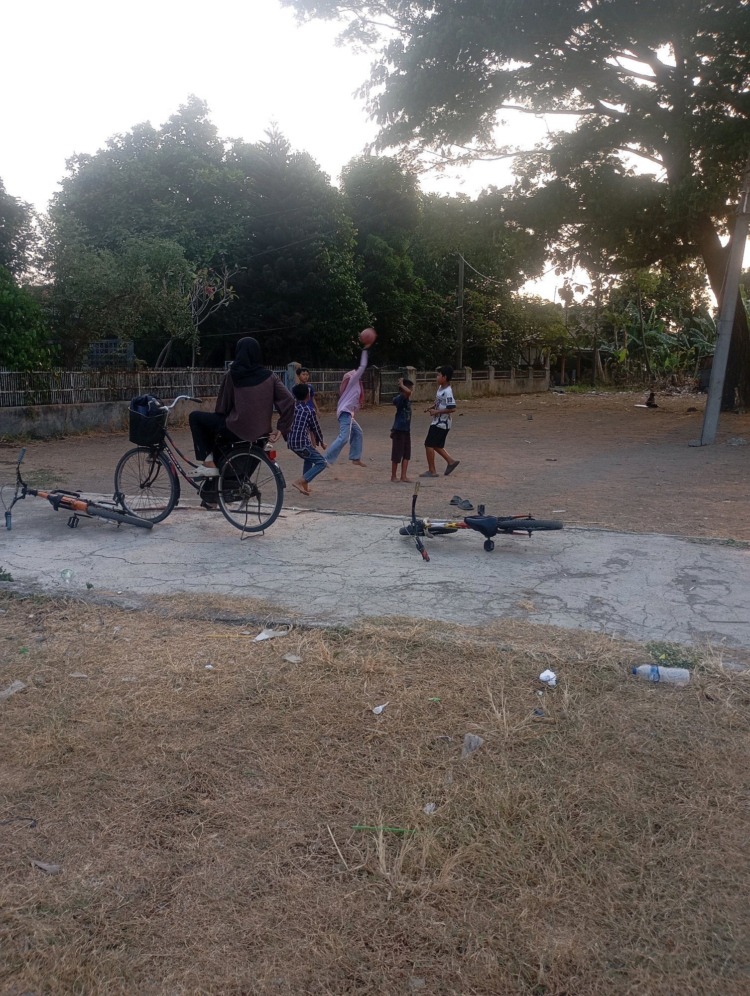 In Babelan, Indonesia, a group of individuals are engaged in a recreational activity in an outdoor, dusty area. A woman wearing a hijab is seated on a bicycle, parked on a concrete surface, while a few children play nearby. One child is in mid-air, jumping, and appears to be reaching for a basketball that is visible in the frame. Another child is standing, watching, and a third child is in the process of walking away. Two bicycles are present; one is upright and ridden by the woman, while the other lies on its side in the foreground. The environment is characterized by dry, sparse grass, a dirt path, and large trees in the background. The lighting suggests it is either early morning or late afternoon.