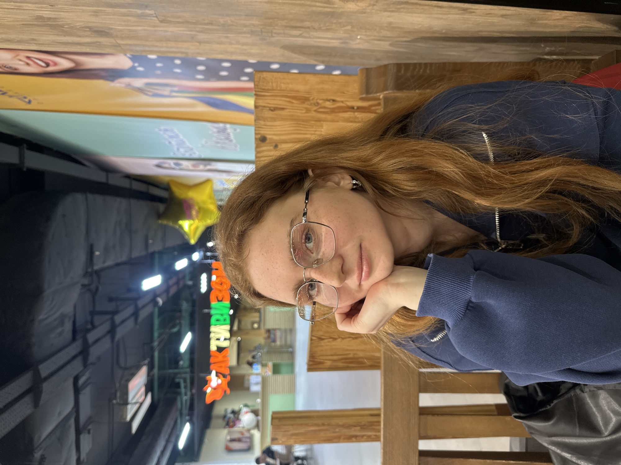 A young, light-skinned woman with red hair and freckles wearing glasses and a dark blue zip-up sweatshirt is seated at a wooden table. Her left hand is supporting her face. Behind her, there is a view of the interior of a public space, possibly a food court or shopping mall. Visible in the background is a sign in Russian, which reads "AK TYMBAL," along with other obscured text. A yellow star-shaped balloon is also visible in the background. The woman's location can be determined as Kazan, Russia based on the sign and other background details.