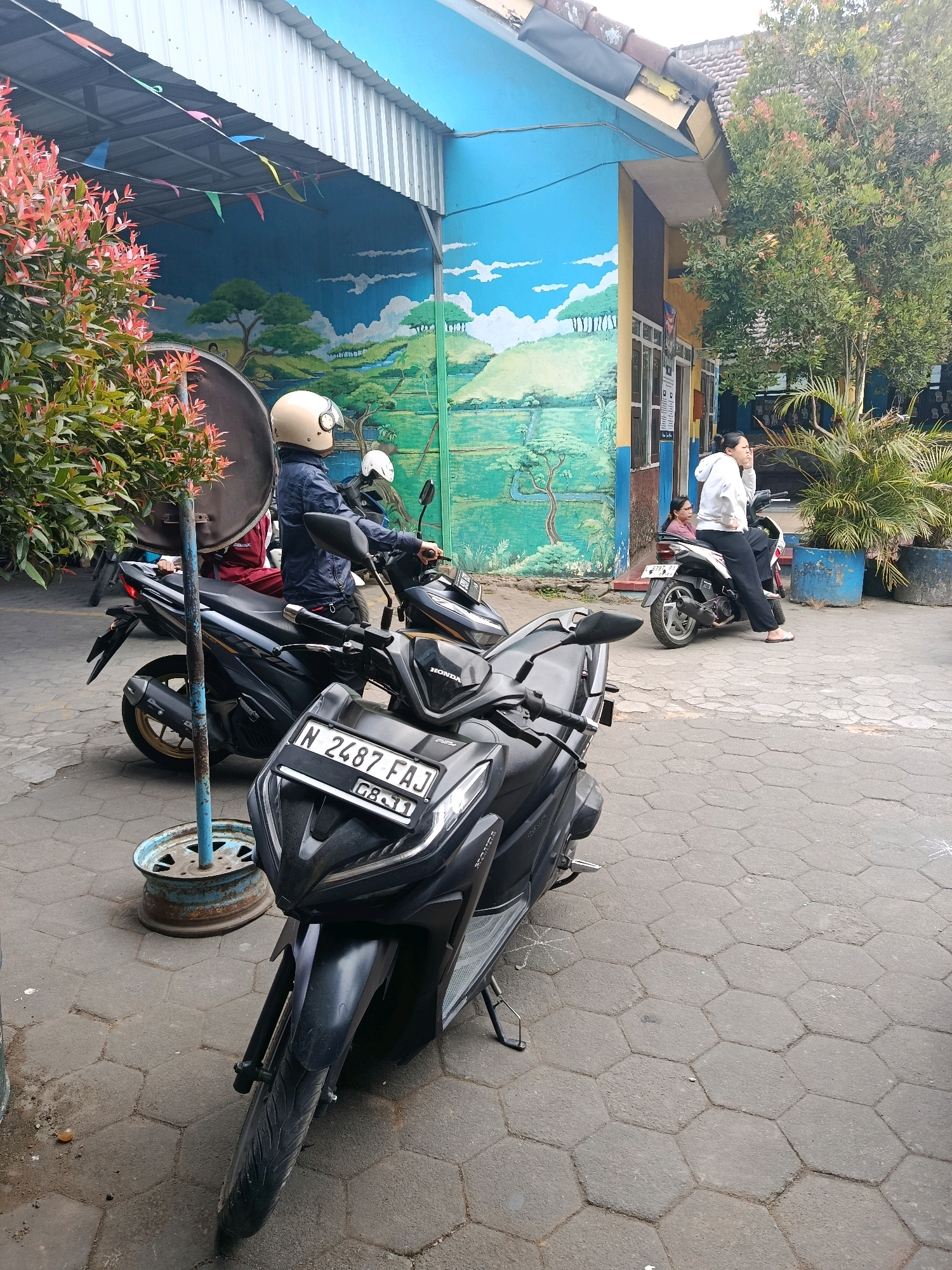 In Bululawang, Indonesia, a street scene unfolds with several parked motorcycles and two individuals. In the foreground, a black Honda motorcycle is prominently displayed, its license plate reading "N 2487 FAJ" and "GB-11". To its left, another black motorcycle is partially visible. Further back, a person wearing a blue jacket and a beige helmet sits on a black motorcycle. Behind them, another motorcycle is parked, with a person in a white hoodie sitting on it, looking towards the right. The background features a building with a vibrant mural depicting a landscape with trees and a river, painted in blues and greens. Red-flowered bushes are on the left, and a large tree is on the right. The ground is paved with dark gray hexagonal stones.