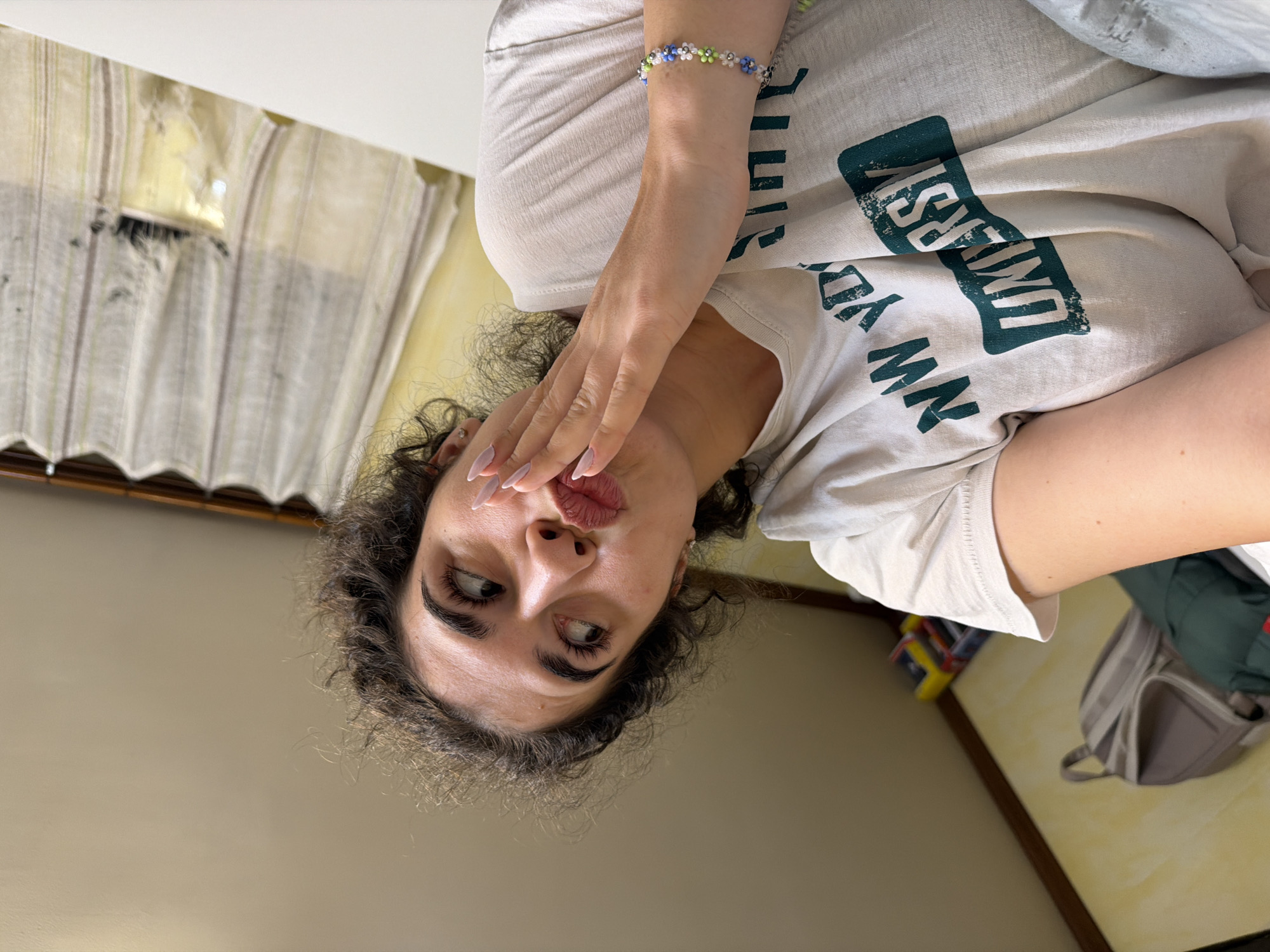 A young woman with curly dark hair is lying down and looking up to her right. She is wearing a light-colored t-shirt with the words "NEW YORK UNIVERSITY" printed on it in green. Her right hand is raised to her face, touching her chin, with her fingers spread. She has long fingernails painted a light pink color. A beaded bracelet with small flower designs is visible on her left wrist. The background consists of a slanted ceiling and a wall with a window covered by sheer curtains. A backpack is visible on the left side of the image. The scene appears to be indoors, possibly in a bedroom or common area.