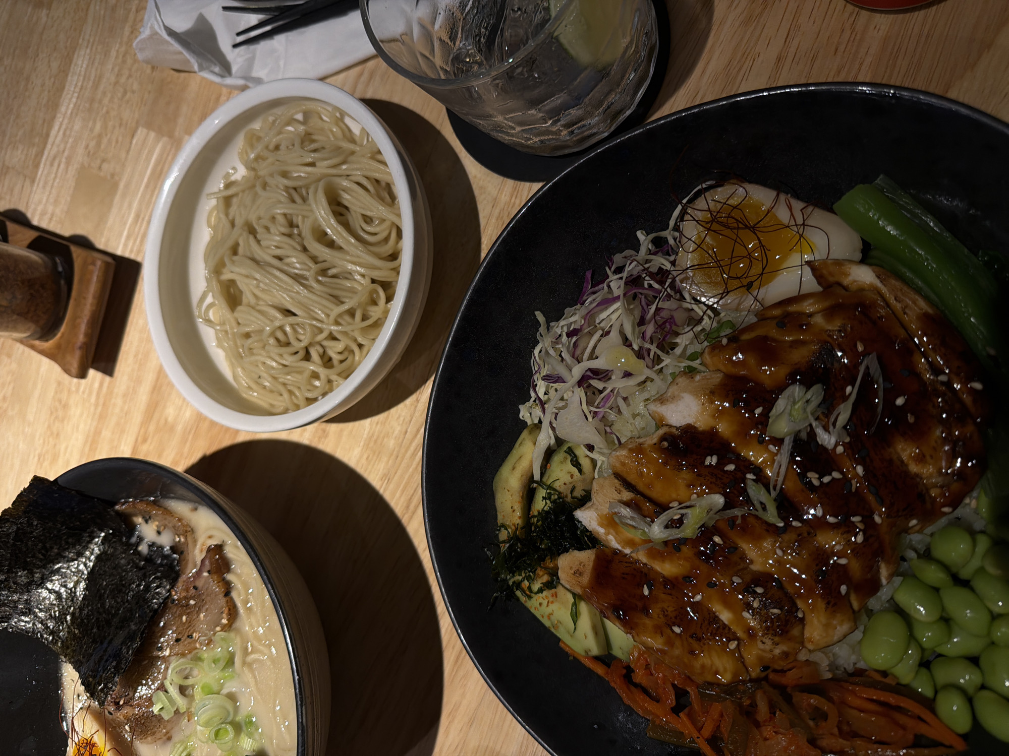 A table is set with a variety of Japanese dishes and a beverage. In the lower left, a bowl of ramen is adorned with seaweed and green onions. To the left of the ramen is a bowl of plain noodles. To the right of the noodles is a glass with a lime wedge. To the right, a black bowl contains a chicken dish with a variety of vegetables, including edamame, carrots, and cabbage, as well as a soft-boiled egg. The scene is set on a wooden table.
