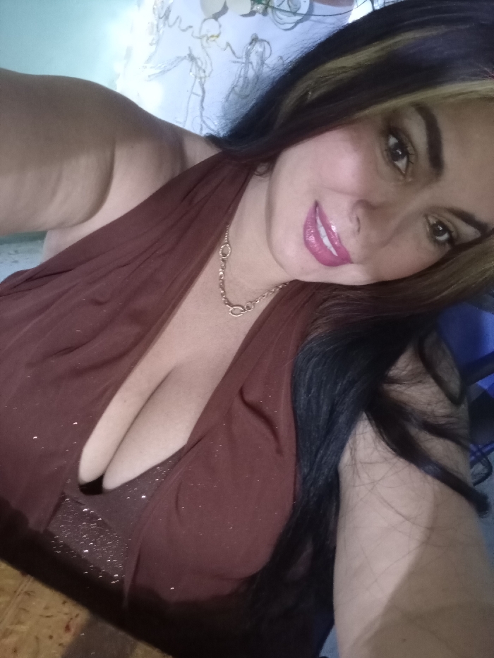 A woman poses for a selfie, her head tilted slightly. She wears a brown, halter-style top that reveals a bit of cleavage. A gold chain necklace adorns her neck. Her dark hair is streaked with blonde highlights. She's wearing lipstick and her skin appears smooth. The background is blurry but features a patterned wall and a hint of a blue object.