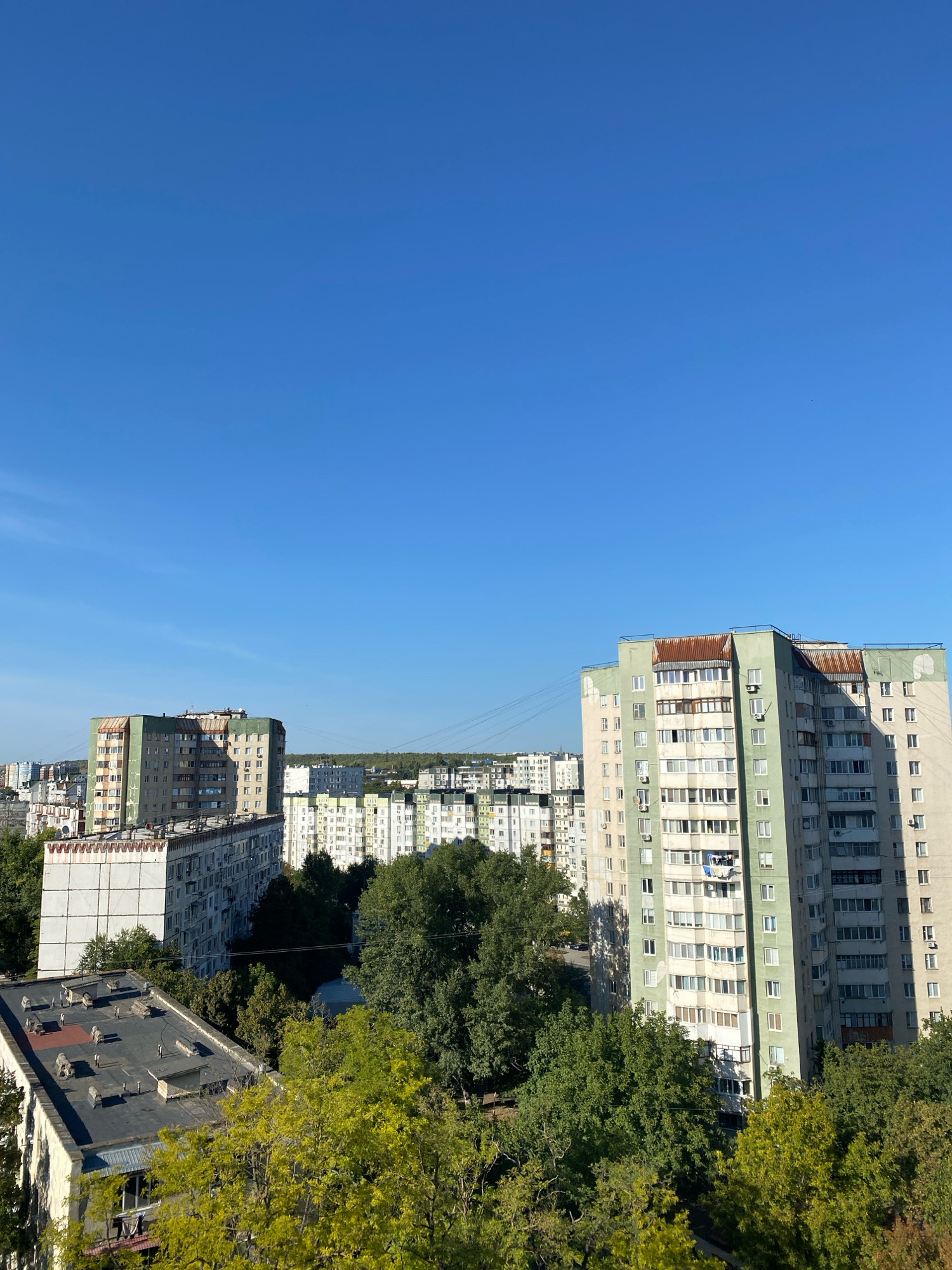The image captures an urban landscape under a clear blue sky, likely taken from a high vantage point in Chișinău, Moldova. In the foreground, the flat, dark roof of a residential building is visible, adorned with various protrusions and vents, and a smaller section of lighter-colored roofing. Beyond this, a cluster of mature trees with vibrant green and yellow foliage forms a dense canopy.
Further into the scene, several multi-story apartment buildings dominate the mid-ground and background. The building closest to the right is a tall, light-colored structure with green accents and numerous windows, some of which are enclosed by balconies. The roof of this building features a distinctive, rusted metal covering. To its left, another apartment building, partially obscured by trees, appears similar in style. In the distance, a panorama of more apartment blocks, predominantly white and light-colored, stretches across the horizon, interspersed with more trees and the faint lines of utility wires. The overall impression is of a typical Eastern European residential district during daylight hours, likely late summer or early autumn given the foliage colors.