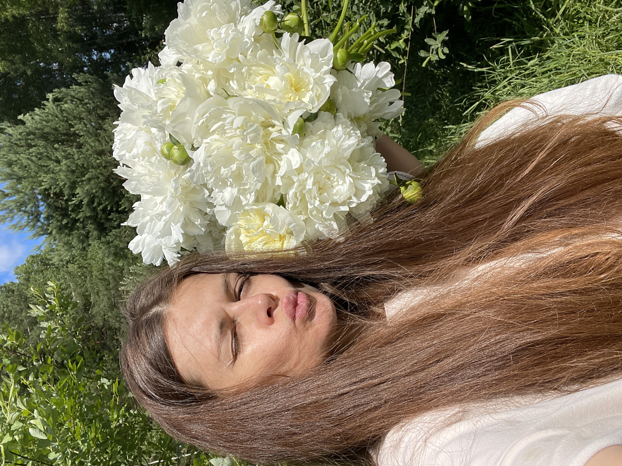 A woman with long, brown hair is taking a selfie outdoors, holding a large bouquet of white peonies. She is wearing a white t-shirt. Her left eye is closed, and her lips are pursed. The background consists of lush green trees and foliage, suggesting a natural, outdoor setting possibly in the city of Zarechny, Russia, given the provided context. The sunlight indicates it is daytime.