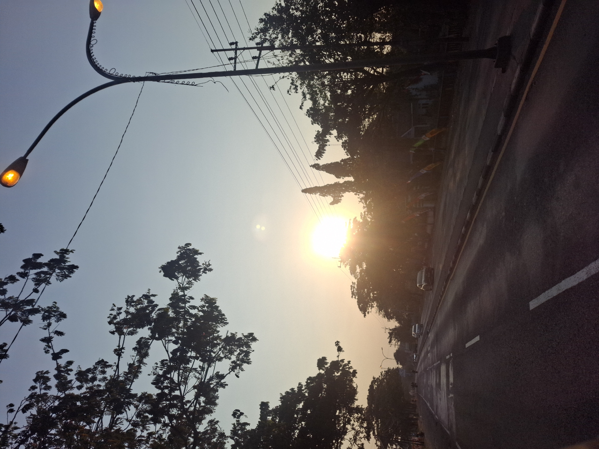 The image depicts a street scene in Paji, Indonesia, during what appears to be dusk or dawn. The sun is low in the sky, casting a bright, warm glow that silhouettes the trees and buildings on either side of the road. A paved road runs through the center of the image, with white lane markings. Several cars are visible on the road, and a motorcycle can be seen in the distance. Streetlights with glowing orange bulbs are present, indicating the approaching night. Power lines and poles are strung along the side of the road. The overall atmosphere is peaceful and serene, with the natural light of the setting or rising sun dominating the scene.