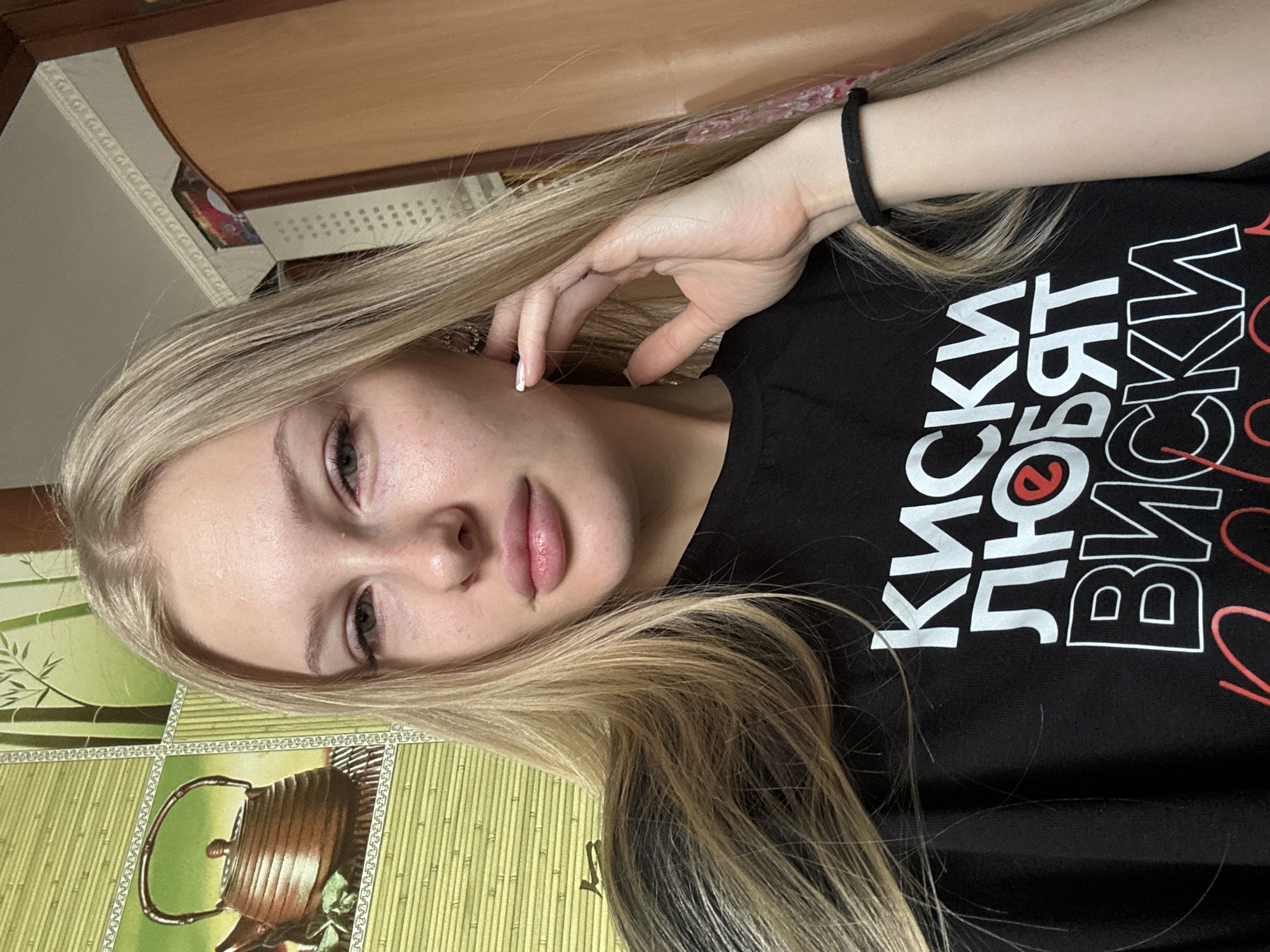 A young woman with blonde hair and light skin is shown from the chest up, looking at the camera. She is wearing a black t-shirt with white text that appears to be in Cyrillic. Her right hand is touching her face. The background is a wall with a green bamboo-themed tapestry on the left and a wooden wardrobe on the right. The image appears to be taken indoors in a residential setting, possibly in Комарихинский, Russia.