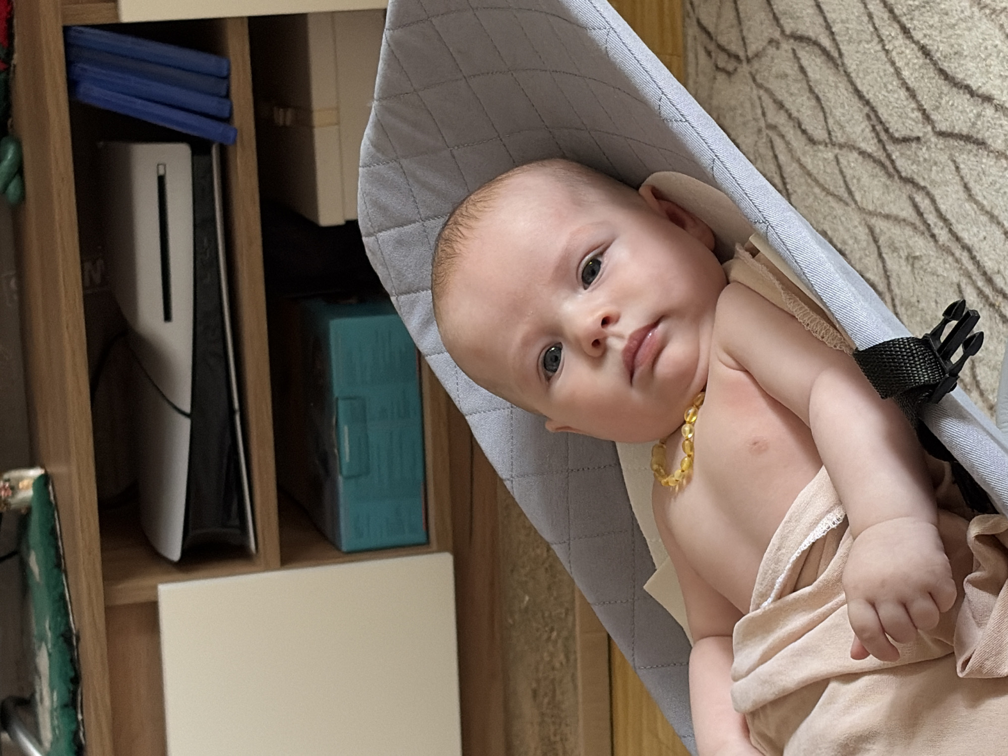 A baby with a light complexion, wearing an amber necklace, lies in a gray baby bouncer. The baby is partially covered with a light beige cloth. To the left, a wooden shelf unit contains a white PlayStation 5 console, blue game cases, and a teal box. The scene is located in Chaikovsky, Russia. The environment includes a beige carpet visible in the upper right.