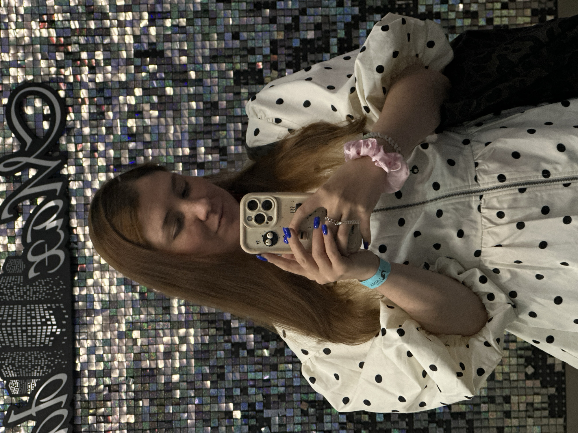 A young woman with long, reddish-brown hair is taking a selfie in front of a shimmering, sequined wall. She is wearing a white dress with black polka dots, puff sleeves, and a zippered front. On her left wrist, she wears a light blue wristband with text on it. Her right hand is holding a smartphone with a light-colored case adorned with two googly eyes. She has bright blue nail polish on her fingers and is wearing a ring on her right ring finger. A pink scrunchie is visible on her left wrist. The background wall is composed of numerous small, square, reflective sequins that create a dazzling, textured effect. Above the sequined wall, a black banner with white graphics resembling stylized buildings and script can be seen. The overall impression is a playful and vibrant scene. There is no information to indicate the location is Petropavlovsk-Kamchatsky, Russia.