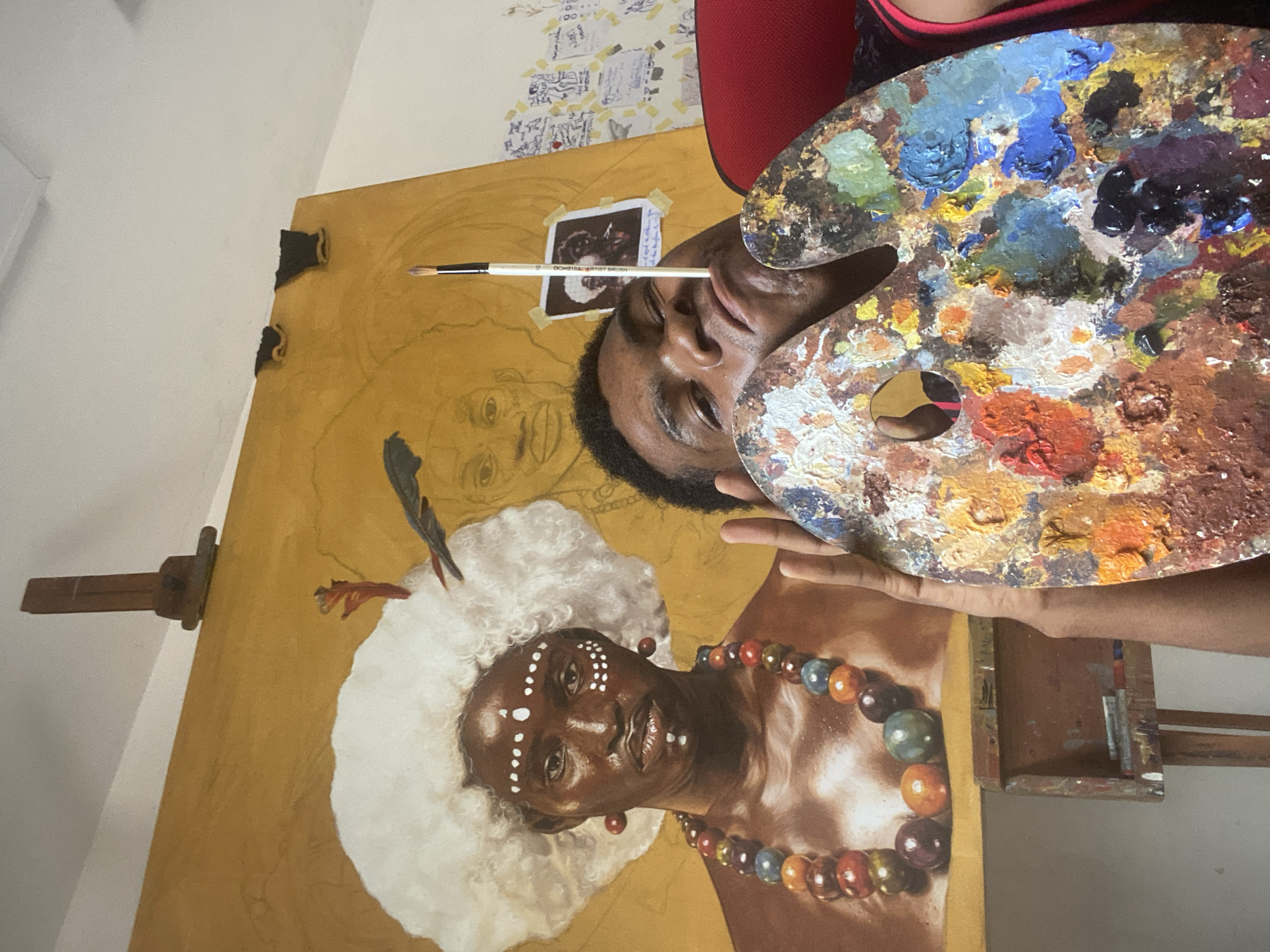 An artist is captured in a close-up, working on a painting, likely in Ado, Nigeria. The male artist, who is Black, is visible from the chest upwards, looking intently at his work. He holds a well-used wooden palette covered with a vibrant array of paint colors, including blues, greens, reds, yellows, and browns, in his left hand. A paintbrush, labeled "DOMESA ARTIST BRUSH" with a visible size '9', is held between his lips.
The large canvas on an easel dominates the scene, featuring a partially completed portrait of a dark-skinned individual, likely a woman, with striking white body or face paint in a pattern of dots. She wears a voluminous, fluffy white headdress or wig and a colorful beaded necklace made of large, varied-colored spheres. A dark feather-like embellishment is visible near her head. The painting's background is a warm golden-yellow, and an outline of another face is faintly visible higher up on the canvas, suggesting ongoing work or a layered composition.
A small, polaroid-style reference photo is taped to the canvas with adhesive tape, depicting a similar figure and aiding the artist. Partially legible handwritten text on the photo's border includes "Love it..." and possibly "100". In the background, a white wall is adorned with several small papers or notes taped to it, and a red object, possibly part of a chair or clothing, is seen behind the artist's head. The wooden easel provides support for the canvas. The overall impression is one of focused artistic creation.