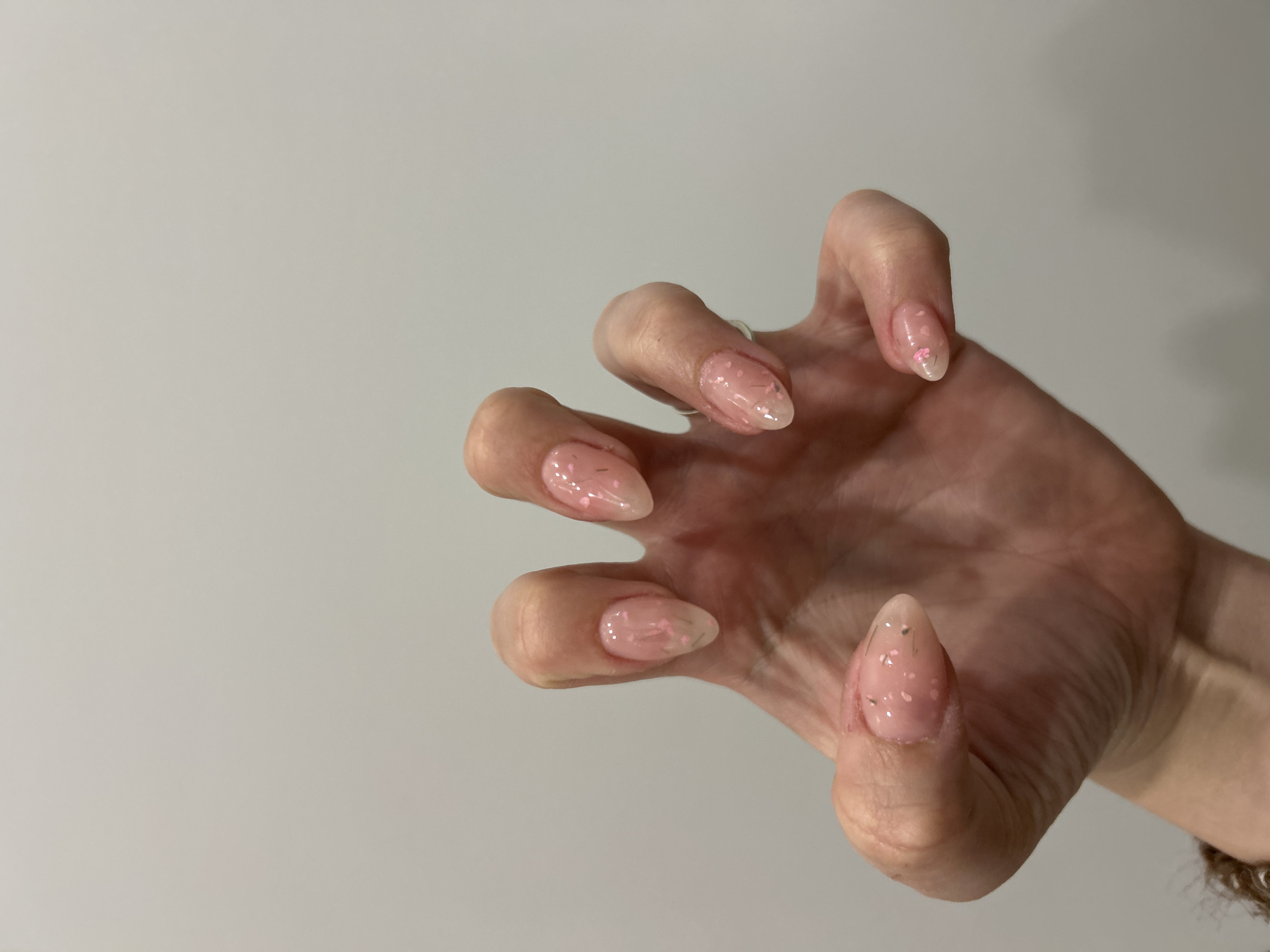 The image presents a close-up view of a person's right hand, likely a woman's, against a plain, light grey background. The hand is positioned with fingers gently curled inward, creating a soft, claw-like or grasping pose, which highlights the manicure. The skin of the hand appears smooth and well-cared-for.
The nails are a central focus, exhibiting a modern almond shape with a translucent, soft pink base. They are adorned with a delicate design consisting of various small, irregularly shaped pieces of pink and iridescent glitter, alongside subtle, tiny black or dark grey linear specks, possibly thin glitter strands, encased within a clear topcoat. These details suggest professional gel or acrylic nail enhancements. A small section of a dark, possibly braided or beaded bracelet or hair tie is visible on the wrist at the bottom right.
The scene is lit evenly and softly, indicative of an indoor environment, making it impossible to determine the time of day or external weather. No specific location cues are present within the image itself, though the context provided states Plovdiv, Bulgaria. There is no visible text. The overall impression is one of a detailed beauty shot intended to showcase the intricate nail art.