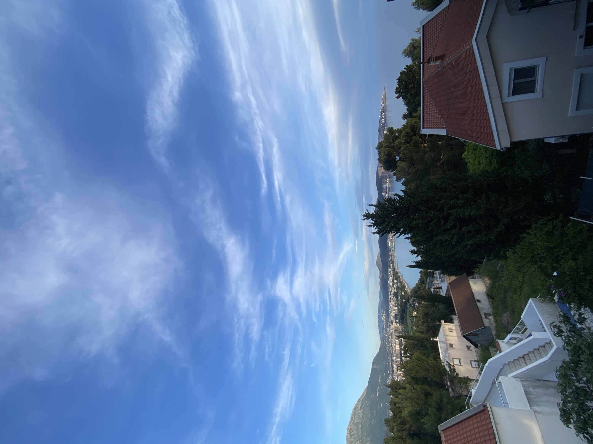 The image shows a view of Šušanj, Montenegro, from an elevated perspective. The sky is blue with elongated, wispy clouds. In the middle ground, there is a cityscape stretching along the coastline, partially obscured by green trees. Mountains are visible in the background, transitioning into the sea. In the foreground are the roofs and sides of residential buildings with red tile roofs and white exteriors. Some buildings have visible windows and staircases. The scene conveys a sunny, residential area with a view of the coast.