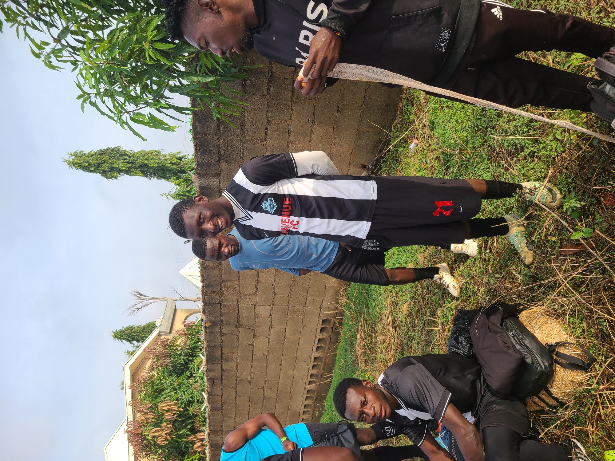 The image depicts four young men in an outdoor setting in Bukuru, Nigeria. One man wears a black Adidas hooded sweatshirt and black pants, holding a white bandage. Another man, in a black and white striped "Avenue FC" jersey and black shorts with the number 21 in red, smiles. A third man wears a light blue shirt and black shorts. A fourth man lies on the ground. A brick wall runs along the background. A house with a white roof is visible in the background, partially obscured by trees. Backpacks and athletic shoes are scattered on the grassy ground, along with twigs and debris.