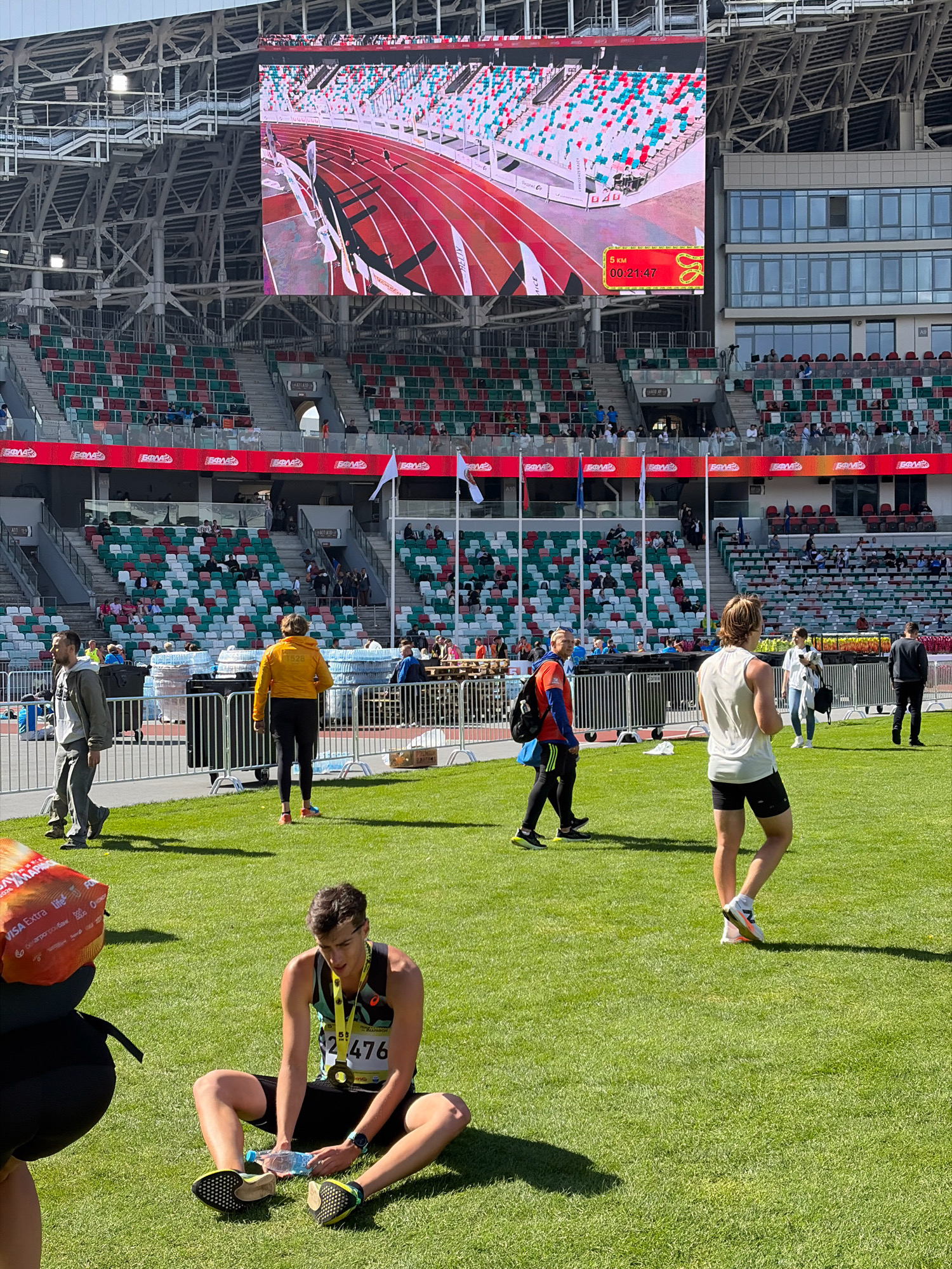A daytime, medium-shot, eye-level view captures a section of the National Olympic Stadium in Minsk, Belarus, during a running event. The stadium features tiered seating in red, white, and green, with a large jumbotron screen displaying a race in progress. In the foreground, a young male runner, wearing a bib numbered 476, a medal, and athletic attire, sits on the grass, holding a water bottle. Other runners and spectators are visible in the background, moving across the field and in the stands. The scene is illuminated by natural sunlight, casting distinct shadows on the green grass.