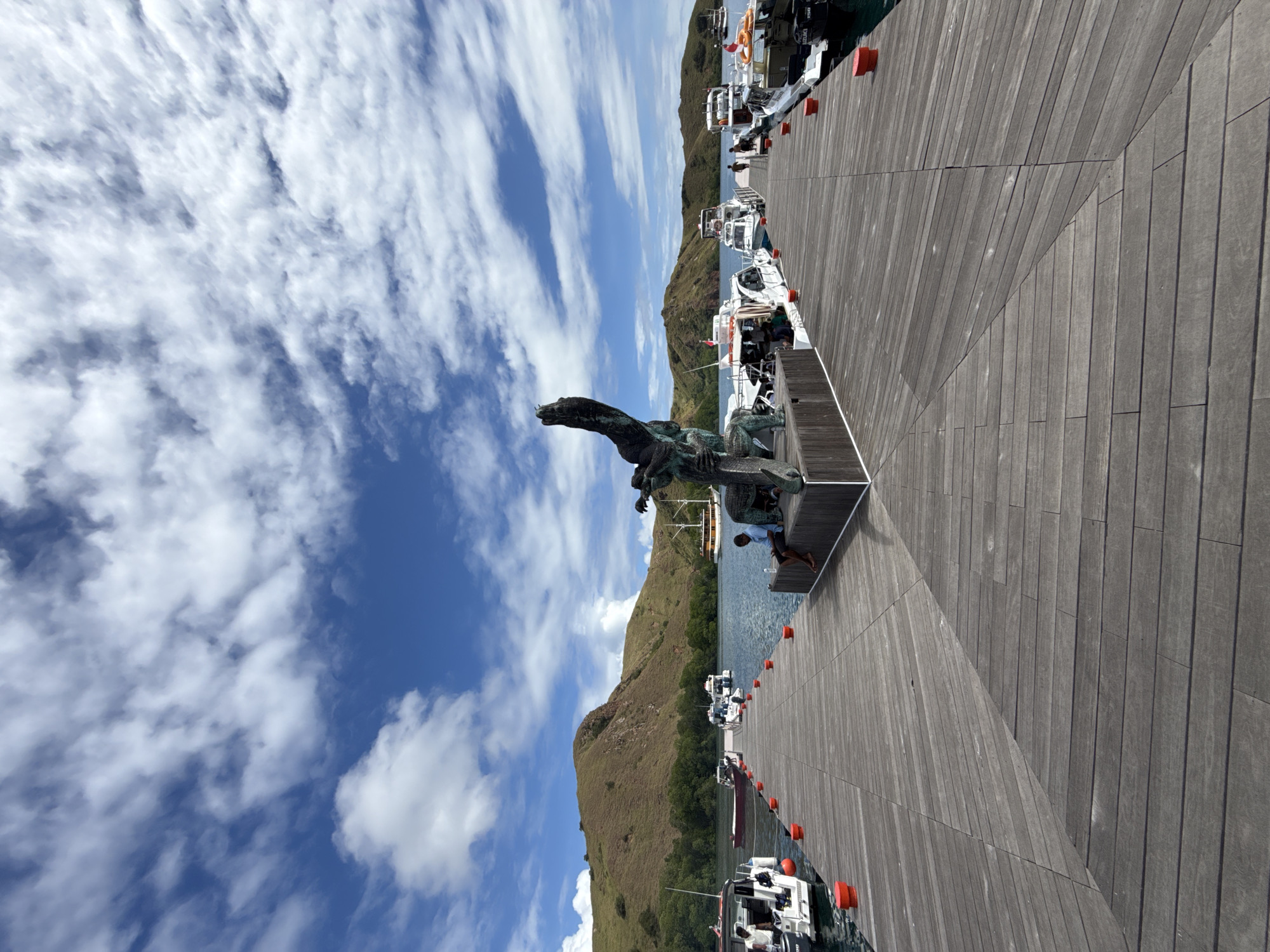 A bronze Komodo dragon statue stands on a wooden platform at a marina in Labuan Bajo, Indonesia. The statue is positioned on the left side of the frame, overlooking the harbor. The marina is lined with docked boats, and a grassy, mountainous terrain can be seen in the background. The sky is blue with scattered white clouds.