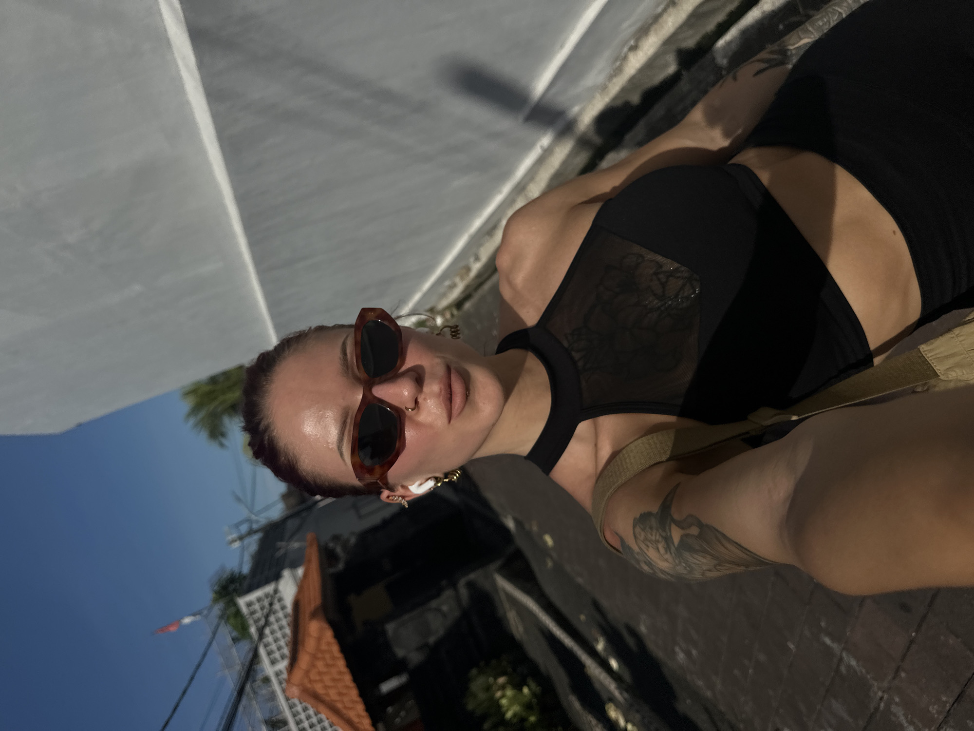 A woman is photographed at an angle, positioned outdoors in North Kuta, Indonesia. She is wearing sunglasses, a black tank top with a mesh panel, and has tattoos on her arm. Her hair is pulled back. The background shows buildings with a red tiled roof, a palm tree, and a partially visible Indonesian flag. The lighting suggests it's daytime, with shadows cast on the pavement and walls surrounding her.