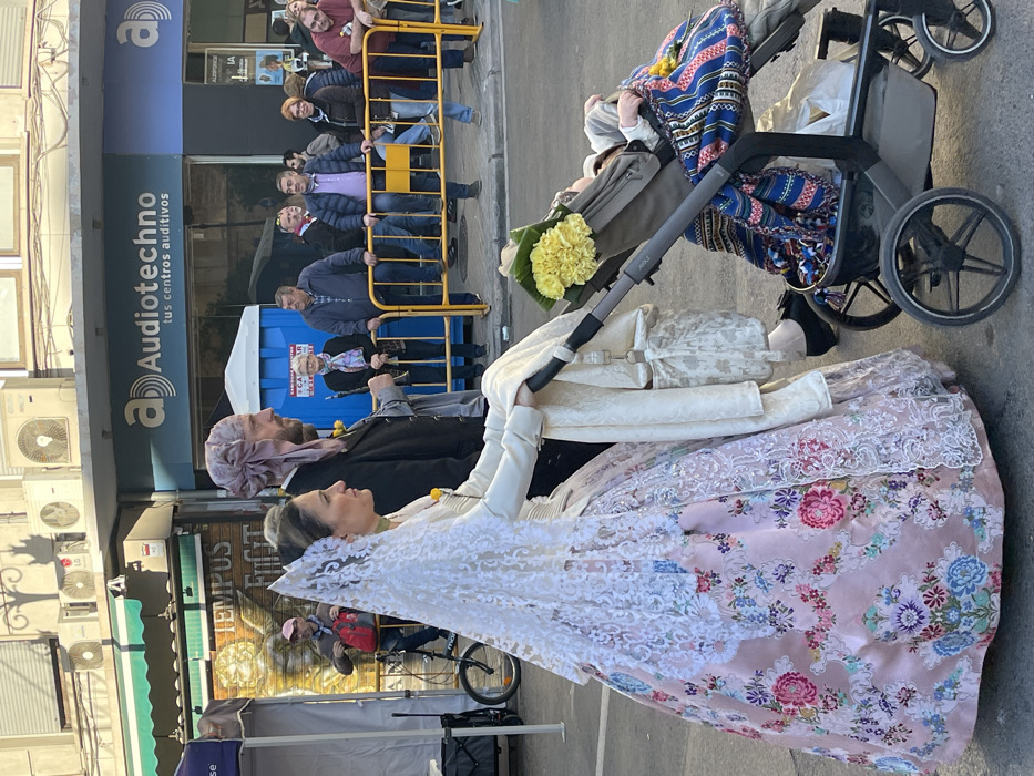 The image depicts a daytime street scene in Valencia, Spain, likely during a cultural event or parade such as the Fallas festival. In the foreground, a woman in traditional Valencian Fallera dress walks on the left, pushing a stroller. Her attire features an elaborate pink silk gown with multi-colored floral patterns, a white lace petticoat, a white lace mantilla-style headdress, a white bodice, and a green choker. Yellow flowers are pinned to her chest. She uses her right hand to push a gray baby stroller containing a baby, partially visible and swaddled in a blue, red, and yellow patterned blanket. Additional yellow flowers are placed on the stroller's canopy and near the baby. A light-colored, patterned bag hangs from the stroller handle.
To the woman's right and slightly behind her, a man in traditional Valencian male attire walks alongside, looking forward. He wears a dark jacket, light-colored undergarments, and a light-colored, possibly silk, head covering. Yellow flowers are pinned to his lapel.
In the midground, a group of spectators observes the scene from behind a yellow metal barrier on the right side of the street. Noteworthy individuals include a man in a blue puffer jacket with his foot resting on the barrier, and an older woman wearing a patterned scarf. Behind the man in traditional dress, a blue portable toilet is visible. Further left, a partial white tent structure is present, and a person on a bicycle wearing a pink cap and red backpack is visible behind it, near a sign reading "TEMPUS FUGIT."
The background features a building with multiple white air conditioning units mounted on its facade. A prominent blue and white sign for "Audiotechno tus centros auditivos" (Audiotechno your hearing centers) is displayed on this building, accompanied by a smaller "AUDIFONOS" sign below it. A dark green awning is visible on an adjacent building to the left. Bright, direct sunlight from the left casts distinct shadows on the ground and buildings.