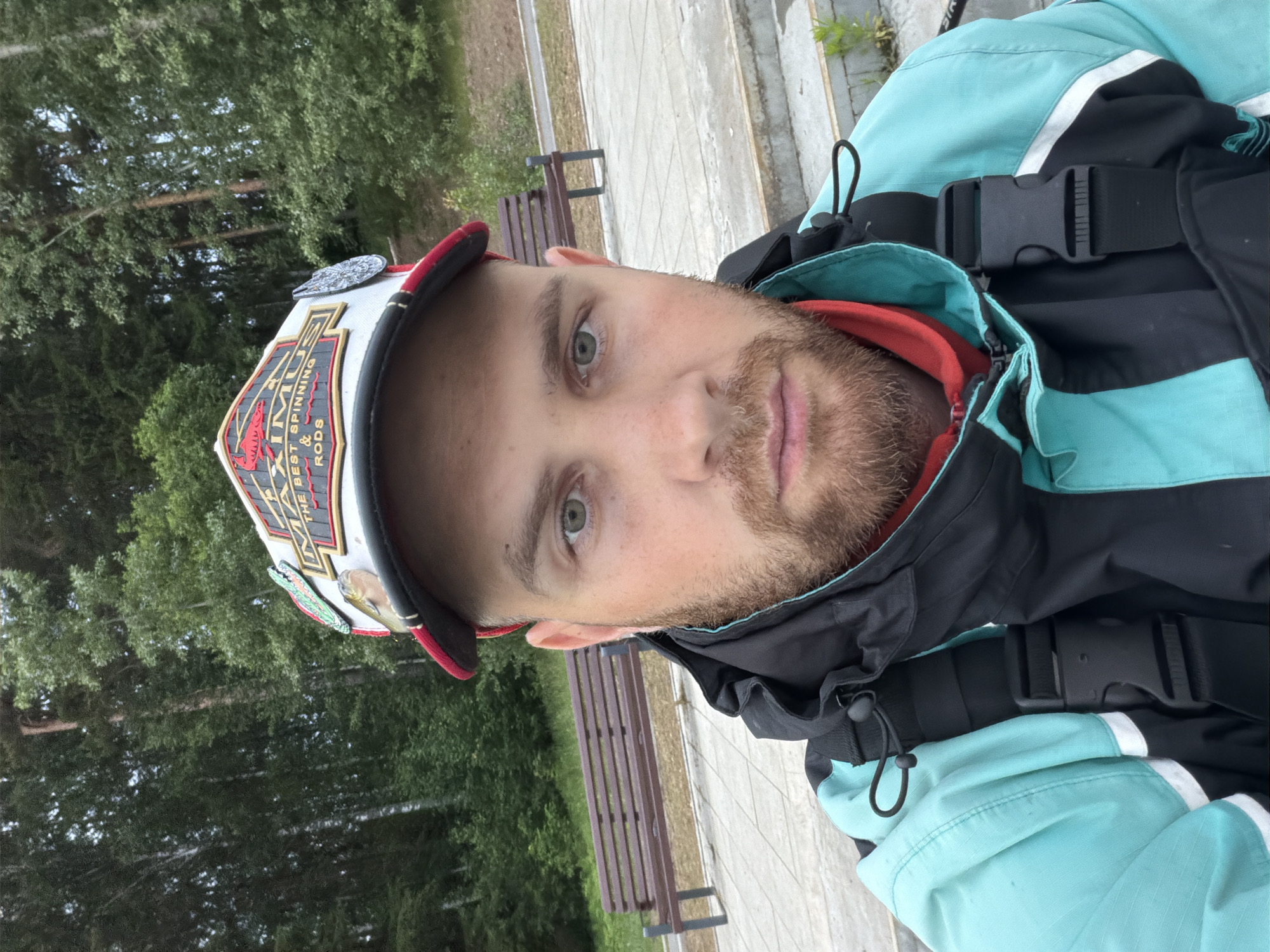 A male individual is depicted in a close-up, head-and-torso selfie, captured outdoors in Chaikovsky, Russia. The subject, a man with fair skin, light-colored eyes, and a light brown beard, wears an off-white and red baseball cap. The cap features a patch reading "MAXIMUS THE BEST SPINNING & RODS" and is adorned with multiple pins, including a green alligator/crocodile, a fish, and a circular metallic emblem. He is dressed in a teal and black jacket with white stripes on the sleeves, a red garment visible at the neck, and a black strap with a plastic buckle, suggesting outdoor or fishing utility wear. The man maintains direct eye contact with the camera, displaying a neutral expression. The background consists of a paved ground surface, a brown wooden park bench to the left, and a dense forest of mixed deciduous and coniferous trees on an upward slope under an overcast sky, indicating a recreational area or park setting.