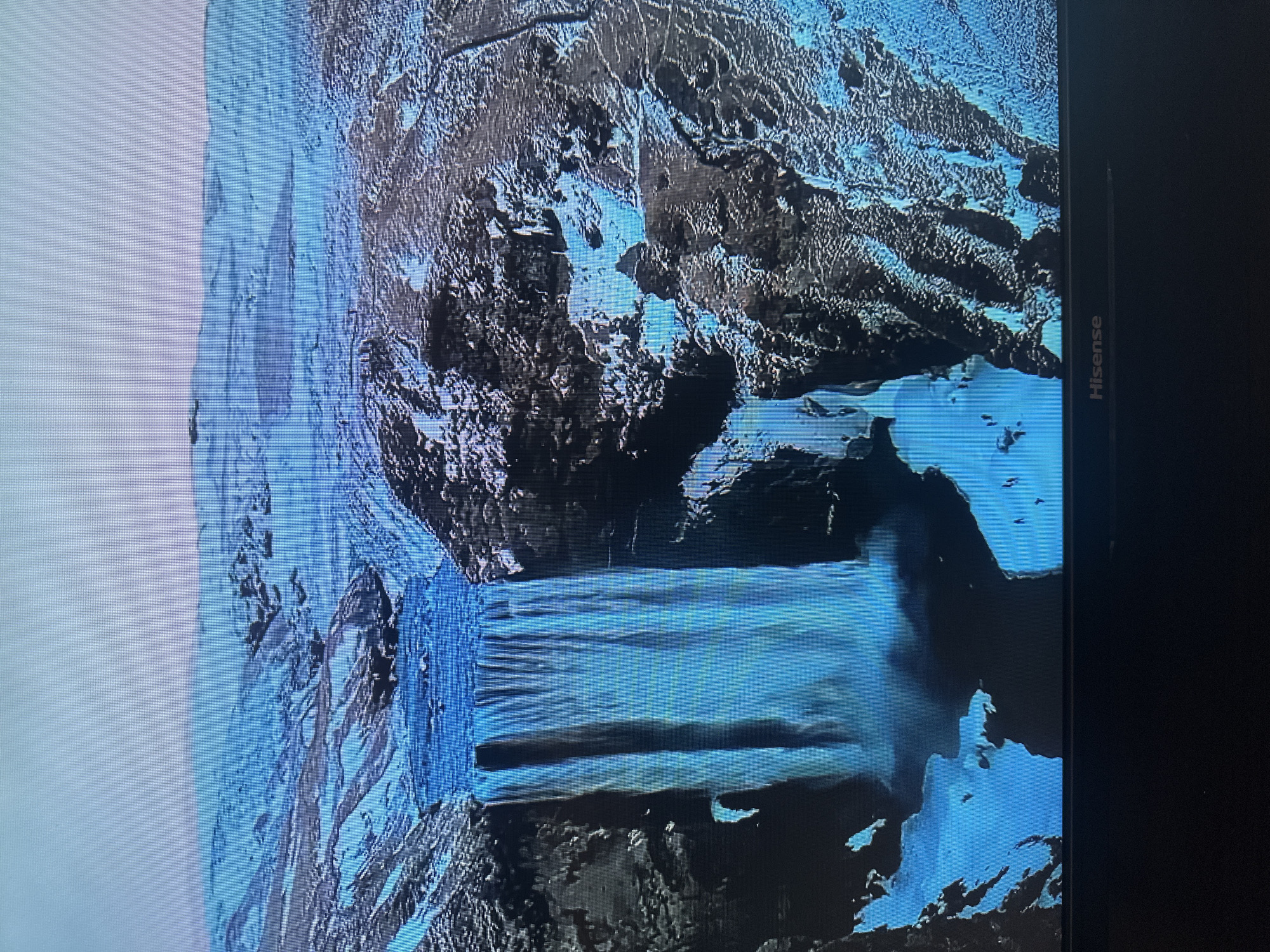 This is an aerial view of a large waterfall cascading over a rocky cliff face. The water is a vibrant blue, and the surrounding landscape is covered in snow and ice, indicating a cold environment. The terrain is rugged and mountainous, with dark rocks contrasting with the white snow and blue water. A narrow river or stream feeds the waterfall from a higher elevation, with hints of a path or road winding along the cliffside in the distance. There are a few small figures visible near the base of the waterfall, suggesting human presence, possibly tourists. The sky is a pale, overcast blue. The image is displayed on a television screen, as evidenced by the visible pixelation and the "Hisense" logo at the bottom. This scene does not depict any location within the city of Kyiv, Ukraine.