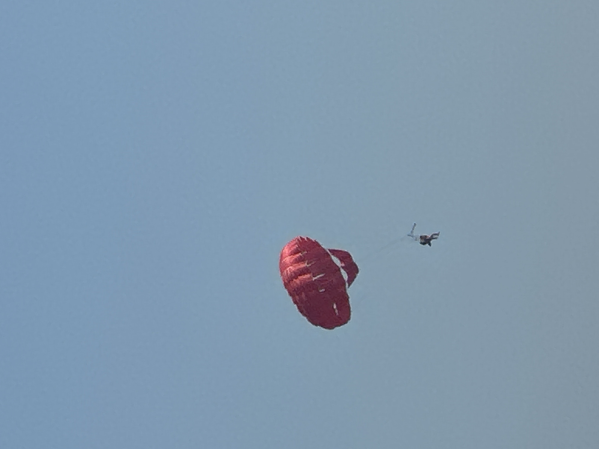 A single person is parasailing in the sky over the city of Boreti, Montenegro. The parasail is red and inflated. The person is suspended below the parasail by ropes and appears to be wearing dark clothing. The sky is a clear blue, indicating it is daytime. There are no other objects or people visible in the image.