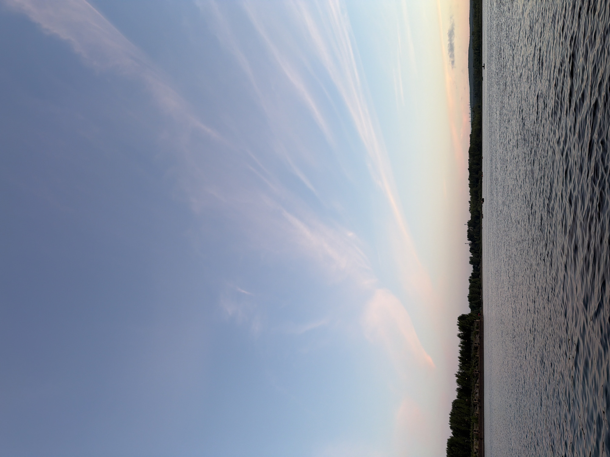 This is an outdoor, wide-angle photograph taken at sunset on a river. The sky is a gradient of pale blue to pink with wispy clouds. The water's surface is rippled and reflects the sky. The far bank is lined with trees, and beyond them, structures suggesting a city or industrial area are visible in the distance. Several small boats are scattered on the water. The scene is tranquil and depicts a natural landscape on the outskirts of Chaikovsky, Russia.