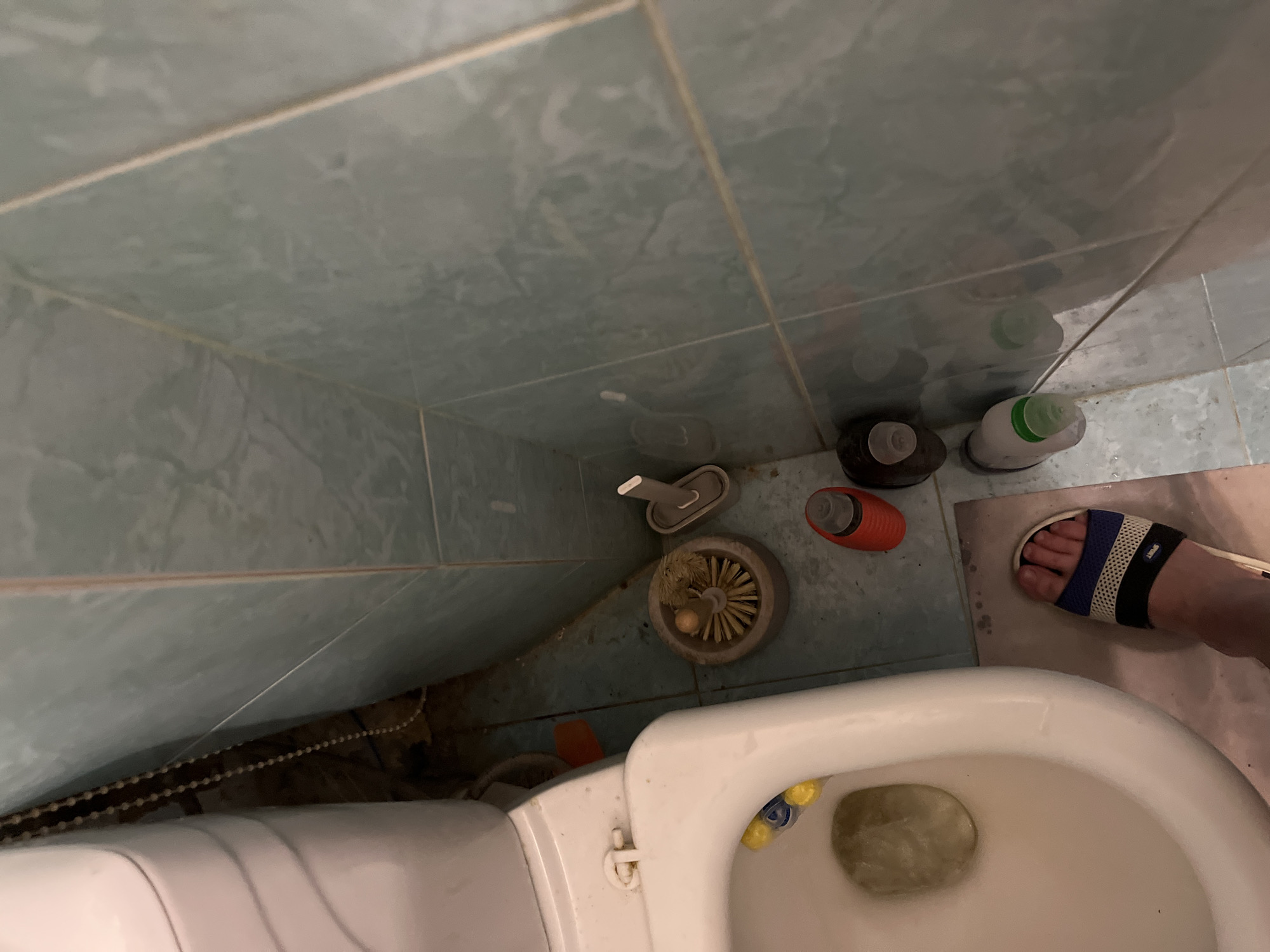The image depicts a high-angle, downward view of a bathroom toilet area in Minsk, Belarus. A Caucasian foot, adorned with a blue, white, and black "SPORT" branded slide sandal, rests on a light-colored bathmat beside a white ceramic toilet. The toilet bowl contains yellowish-brown water, with two yellow and blue cleaner blocks visible within the rim. The toilet lid and seat show some discoloration near the rear hinge. The floor and lower wall are clad in light blue-green tiles, exhibiting noticeable dirt, grime, and discolored grout lines. Various items are on the floor to the right of the toilet: a wooden toilet brush within a circular holder, a gray plastic toilet brush with its holder, a red plastic bottle with a white cap, a dark brown/black bottle with a white cap, and a white/green bottle with a green cap. A white beaded chain hangs behind the toilet tank. A small orange rectangular object is partially visible on the floor behind the toilet brushes. The environment presents signs of routine use and diminished cleanliness.
