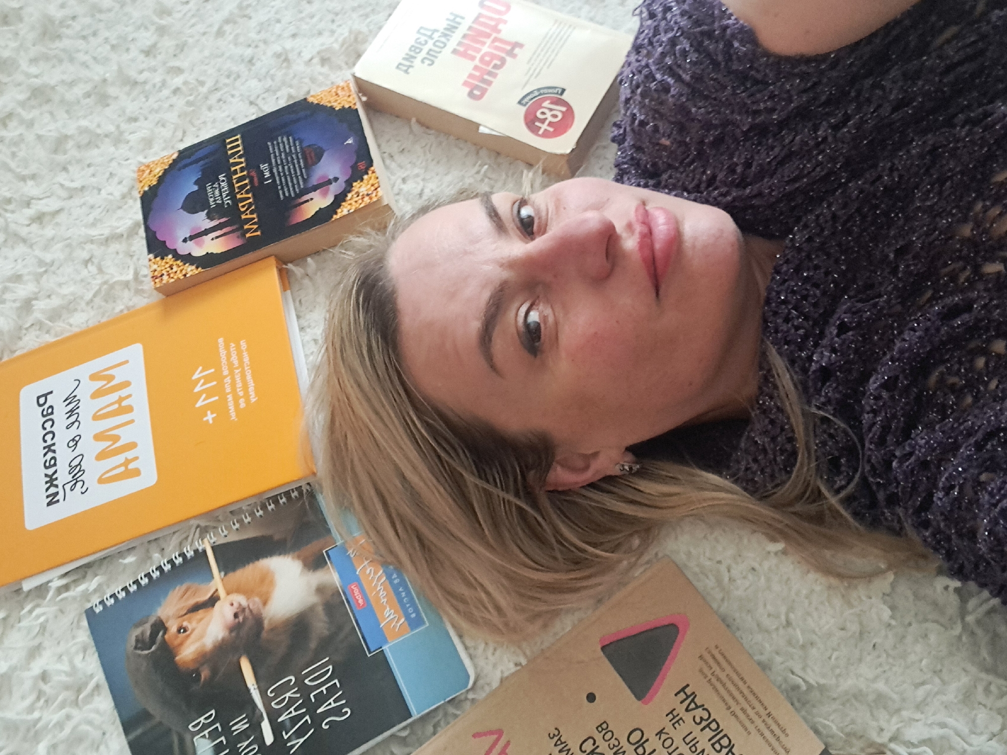 A woman is captured in a close-up, eye-level shot, lying on a plush, light-colored rug, looking directly at the camera. Her head is tilted, and her blonde or light brown hair spreads around her. She wears a dark purple, sparkling, possibly knitted, garment. Her expression is calm and neutral.
She is surrounded by several books and a notebook, suggesting a relaxed reading or creative environment. The books are in various positions, some open, some closed.
Visible texts on the books and notebook include:
- An orange book with "МАМ" (Mom) and "Расскажи" (Tell me) on the spine, and "+777".
- A dark book featuring a silhouette of a mosque or minarets, titled "МАХАТМА".
- A light-colored book, positioned near her head, clearly displaying "ОДИН ДЕНЬ" (ONE DAY) and "ДЭНВЭЛ НИКОЛС" (DANIEL NICHOLLS), along with an "18+" age restriction symbol.
- Another light brown book, partially visible, with "НЕУБИВАЕМОЕ" (INDESTRUCTIBLE) and "НЕ ЦЕ" (NOT THIS), with some other text partially obscured.
- A spiral-bound notebook, featuring an image of a dog holding a paintbrush, with words like "IDEAS" (ИДЕИ) and "Вдохновение" (Inspiration) visible, along with "edford" and "ГОТОВА 84" (READY 84).
The setting appears to be indoors, likely in Elektrostal, Russia, given the common Cyrillic script on the books. The soft rug and collection of reading materials create a cozy atmosphere.