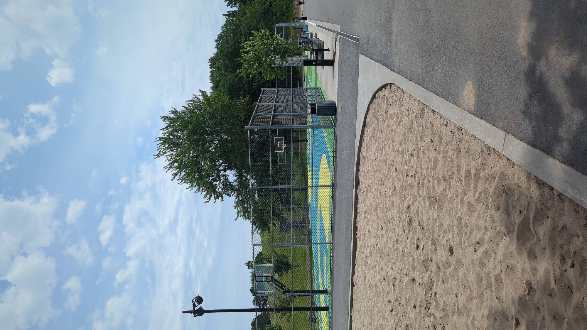 The image depicts an outdoor basketball court and playground area on a cloudy day. In the foreground, a large patch of sand is visible, bordered by a concrete edge and an asphalt pathway. Beyond the sand, a fenced basketball court with a brightly colored surface is present. The court features two basketball hoops. A street lamp stands to the left of the court. In the background, trees and a grassy hill are visible. Several benches and trash receptacles are positioned along the edge of the basketball court. A few individuals are walking on the pathway in the distance. The scene is likely located in an urban park setting within Montreal, Canada, given the presence of various recreational amenities and typical urban park landscaping.
