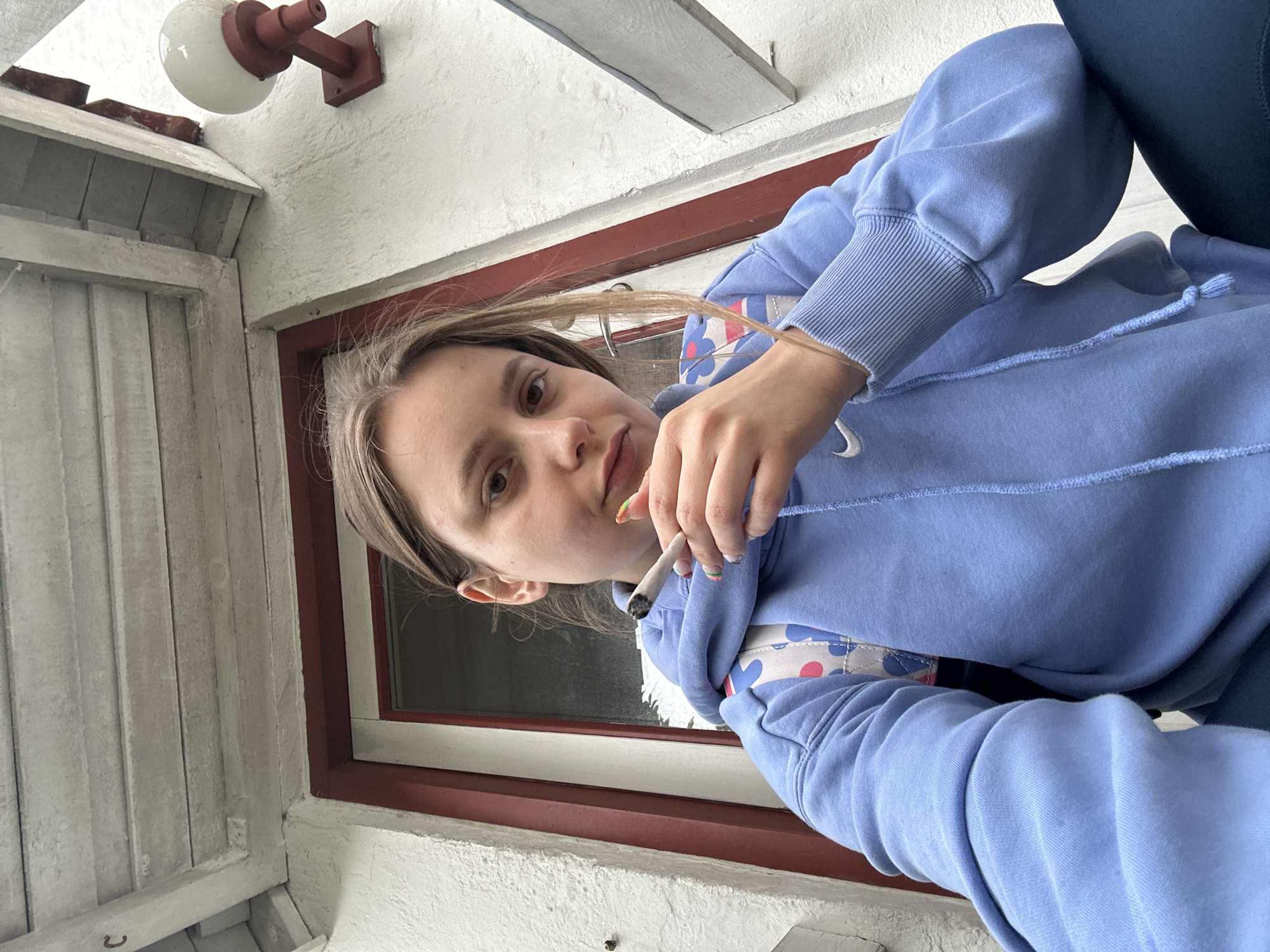 A young woman is depicted in a close-up, low-angle shot, looking directly at the camera with a neutral expression. She has light brown hair, which frames her face. She is wearing a light blue hooded sweatshirt with a drawstring at the neckline and ribbed cuffs; a faint white Nike swoosh is visible on the left cuff. A subtle pattern of light blue, white, and pink shapes is visible on the front of her hoodie. Dark-colored pants are visible on her right side.
In her right hand, she holds a lit cigarette between her thumb and index finger. Her fingernails are painted with distinctive multi-colored tips. The background suggests an outdoor or semi-outdoor setting, possibly a porch or balcony. To her left, white painted wooden siding is visible. Behind her, a window with a prominent dark red frame is seen, showing a dark interior beyond the glass. Above her, a white textured wall features a wall-mounted light fixture with a spherical white globe and a reddish-brown base. The scene appears to be taking place during daylight hours in Horsens, Denmark. No other explicit text is visible within the frame.