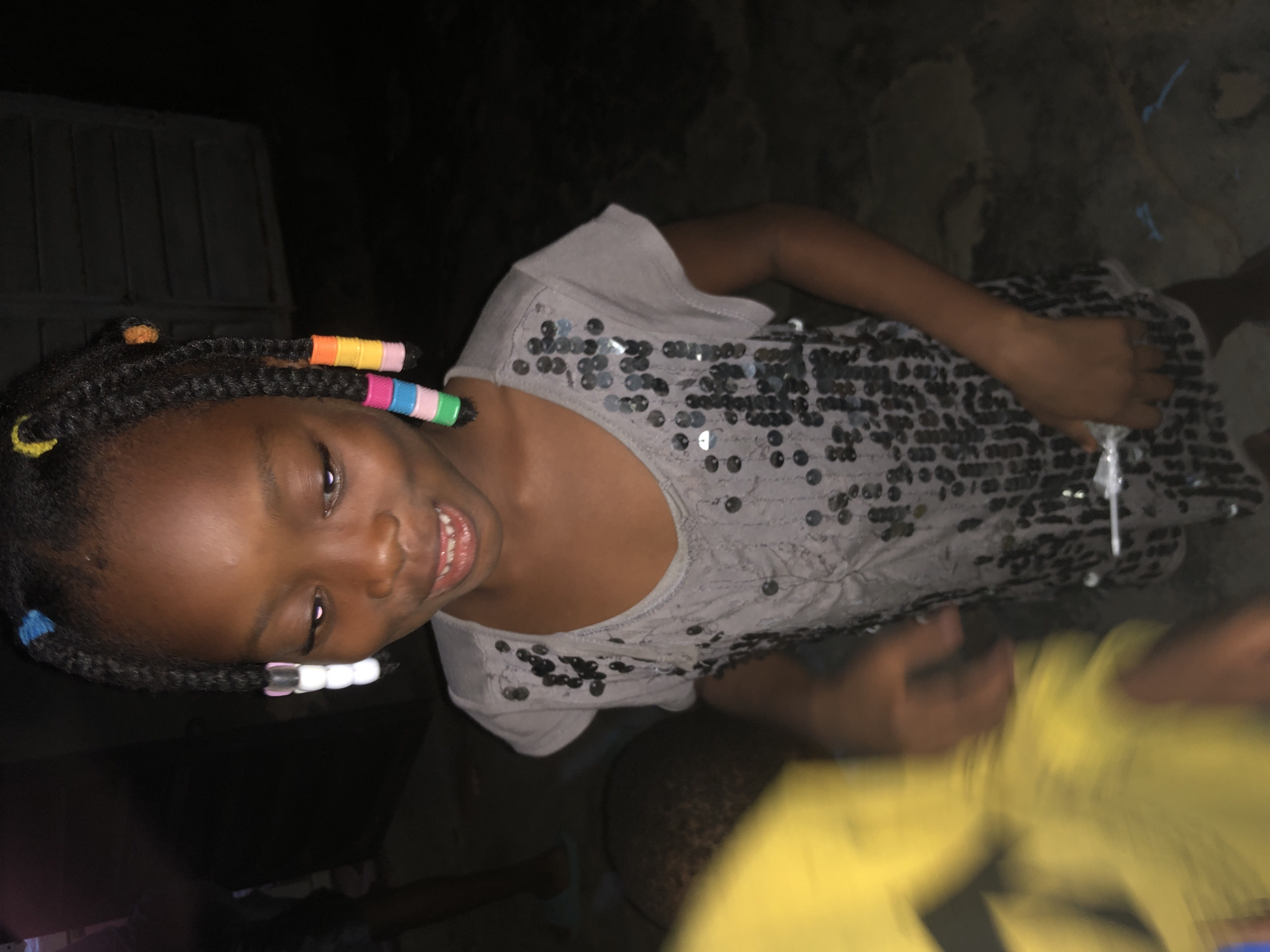 The image captures a young Black girl with braided hair adorned with colorful beads, smiling. She is wearing a light grey t-shirt embellished with numerous dark sequins. The background is dark, suggesting an outdoor setting at night. The girl's skin tone is dark brown. In the lower left portion of the image, a blurry yellow object is visible, possibly another person or an object. The ground appears to be dirt or a rough surface. The scene suggests a casual, possibly festive, gathering. It is not possible to determine the location within the city of Ado, Nigeria from the visual information provided.