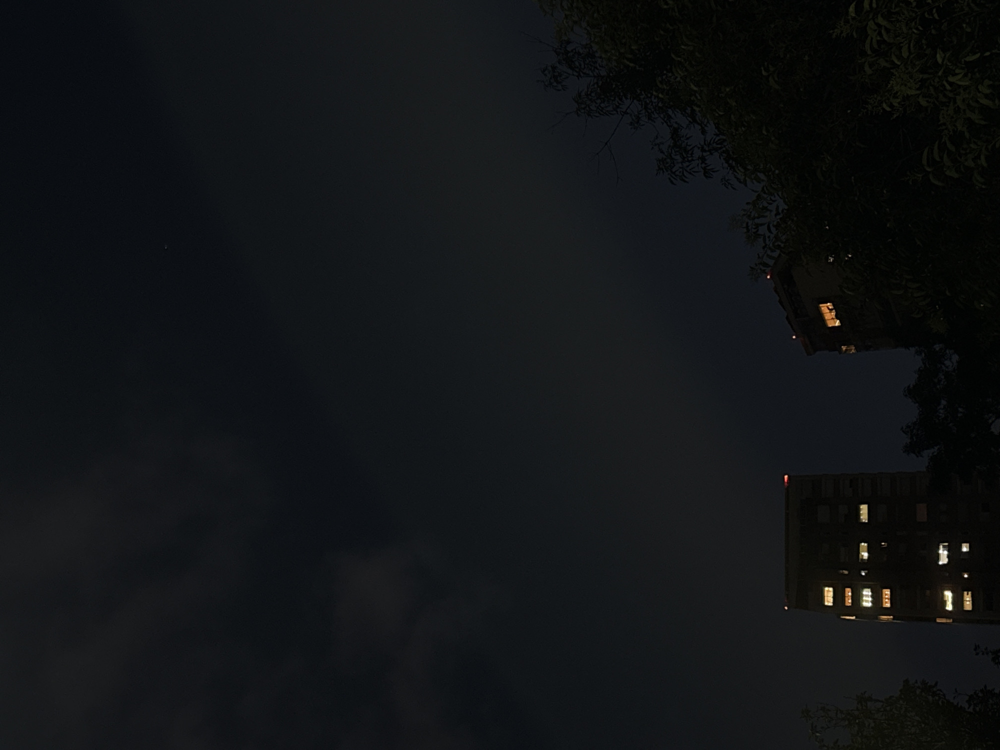 The image shows a night scene in Mexico City, Mexico, with a dark sky and a faint star visible in the upper left. Two buildings are silhouetted against the night, both displaying illuminated windows. Tree foliage partially obscures the upper right corner of the image.