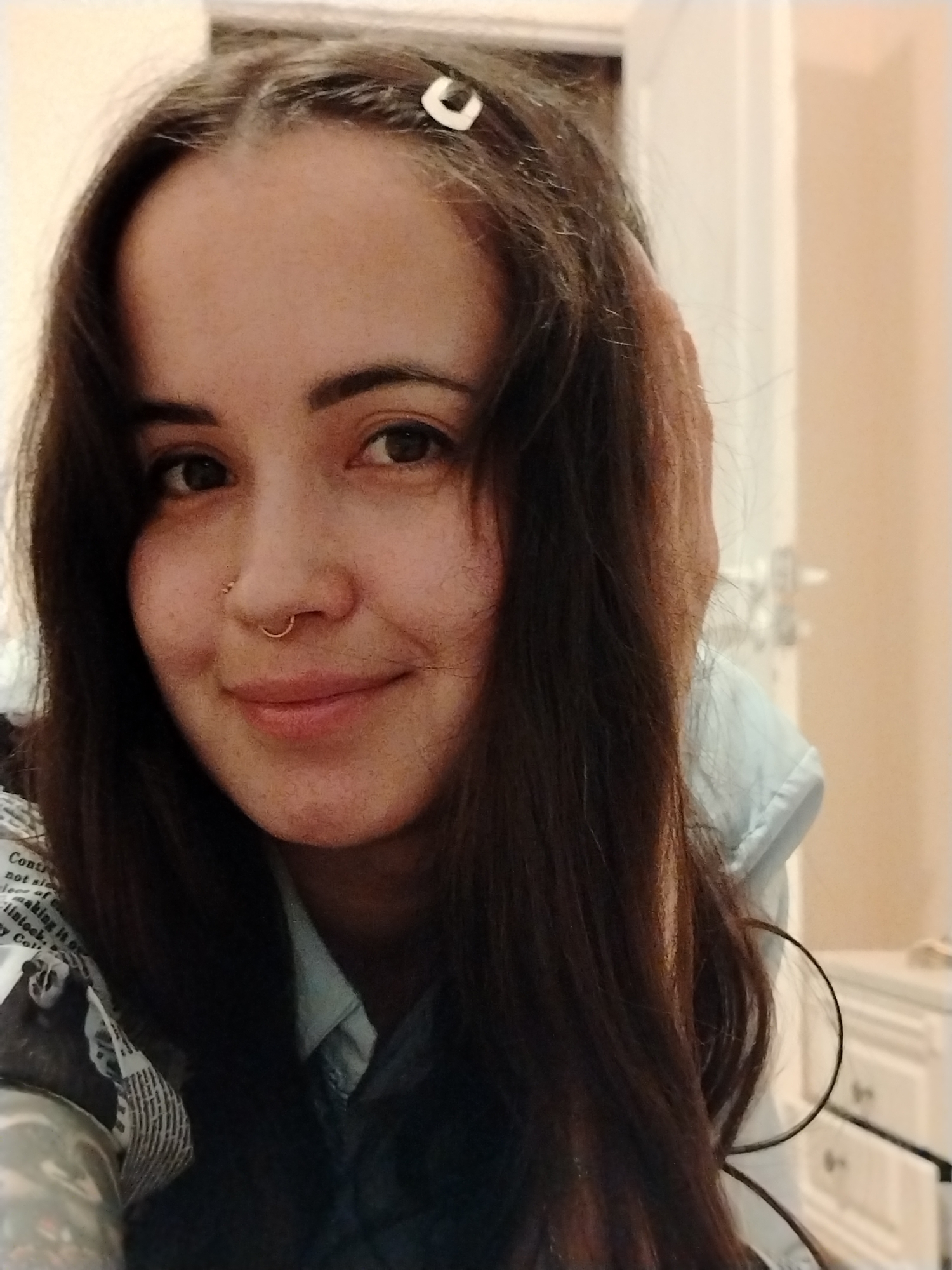 A close-up portrait shows a young woman with long, dark brown hair. She is fair-skinned with brown eyes and a gold nose ring. She has a slight smile. A small, white hair clip is visible in her hair. She wears a light blue collared shirt under a darker jacket printed with newspaper-like text. The background is blurred, showing a white door and furniture, suggesting an indoor setting. There is no indication of being in Noviy, Russia.