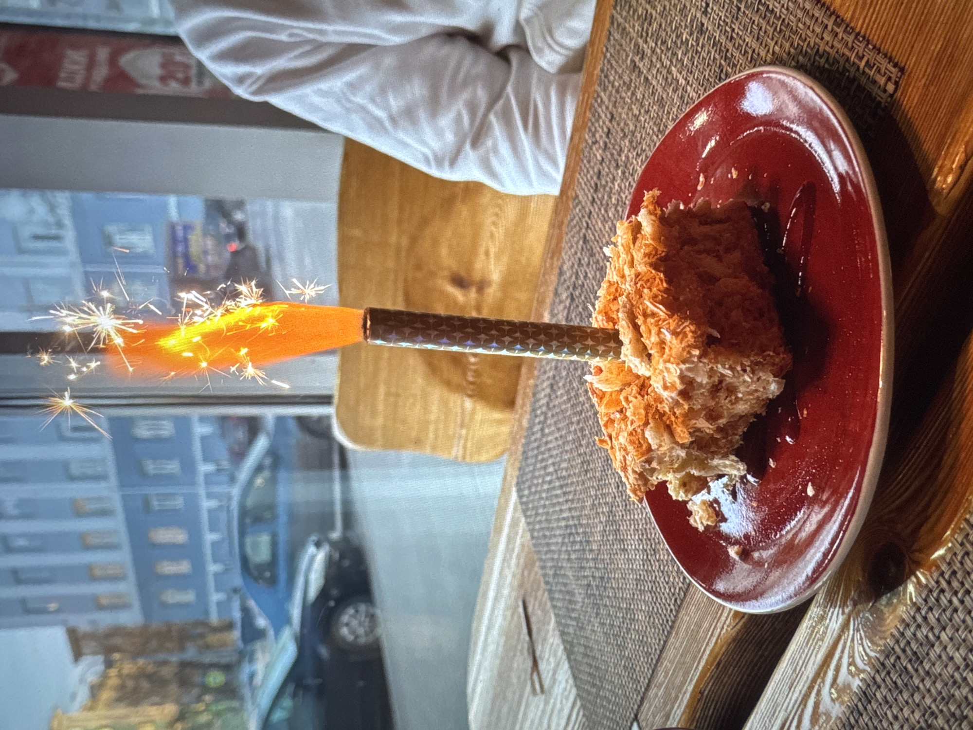 A close-up, low-angle shot shows a slice of Napoleon cake on a red plate. A sparkler is lit and placed in the cake, emitting bright orange flames and numerous white sparks. The cake appears to be topped with shredded coconut or frosting. The plate sits on a woven placemat, which is on a wooden table. In the background, a blurred cityscape is visible through a window. The city appears to be Новинки, Russia, indicated by a blue building and cars on a street. A person's shoulder and arm, clad in a white garment, are visible on the right side of the frame. The overall atmosphere suggests a celebration.