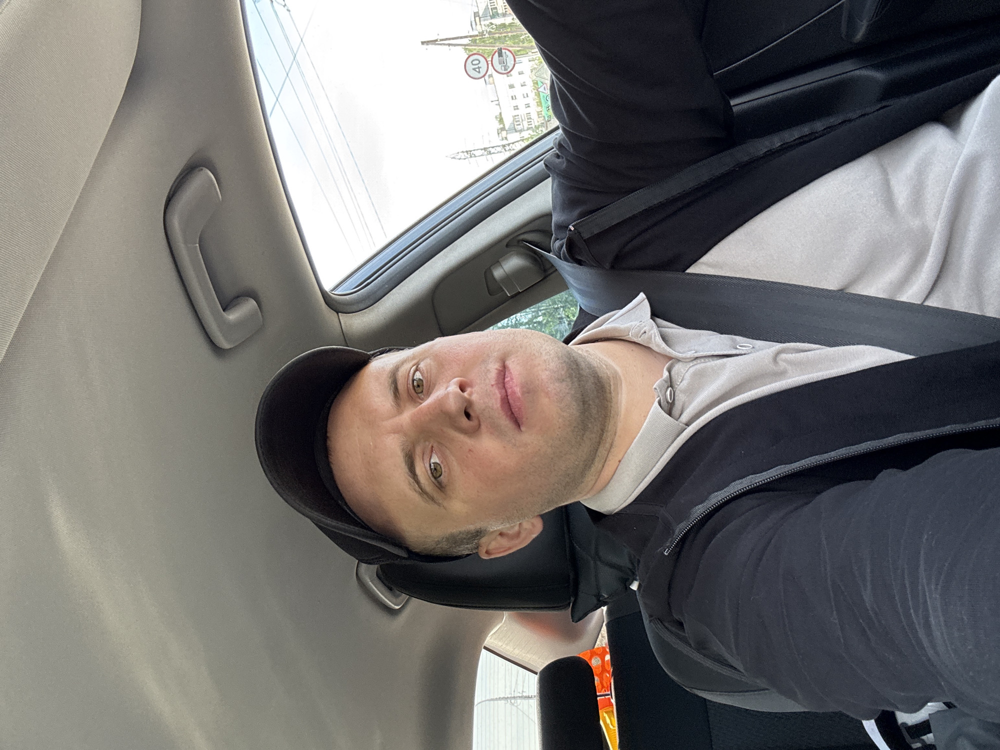 A man wearing a black baseball cap, a beige collared shirt, and a black jacket with a seatbelt is sitting inside a car. The car's interior is gray. Outside the car window, a street sign in the city of Chaikovsky, Russia is visible. The photo appears to be taken from a low angle, looking up towards the man.
