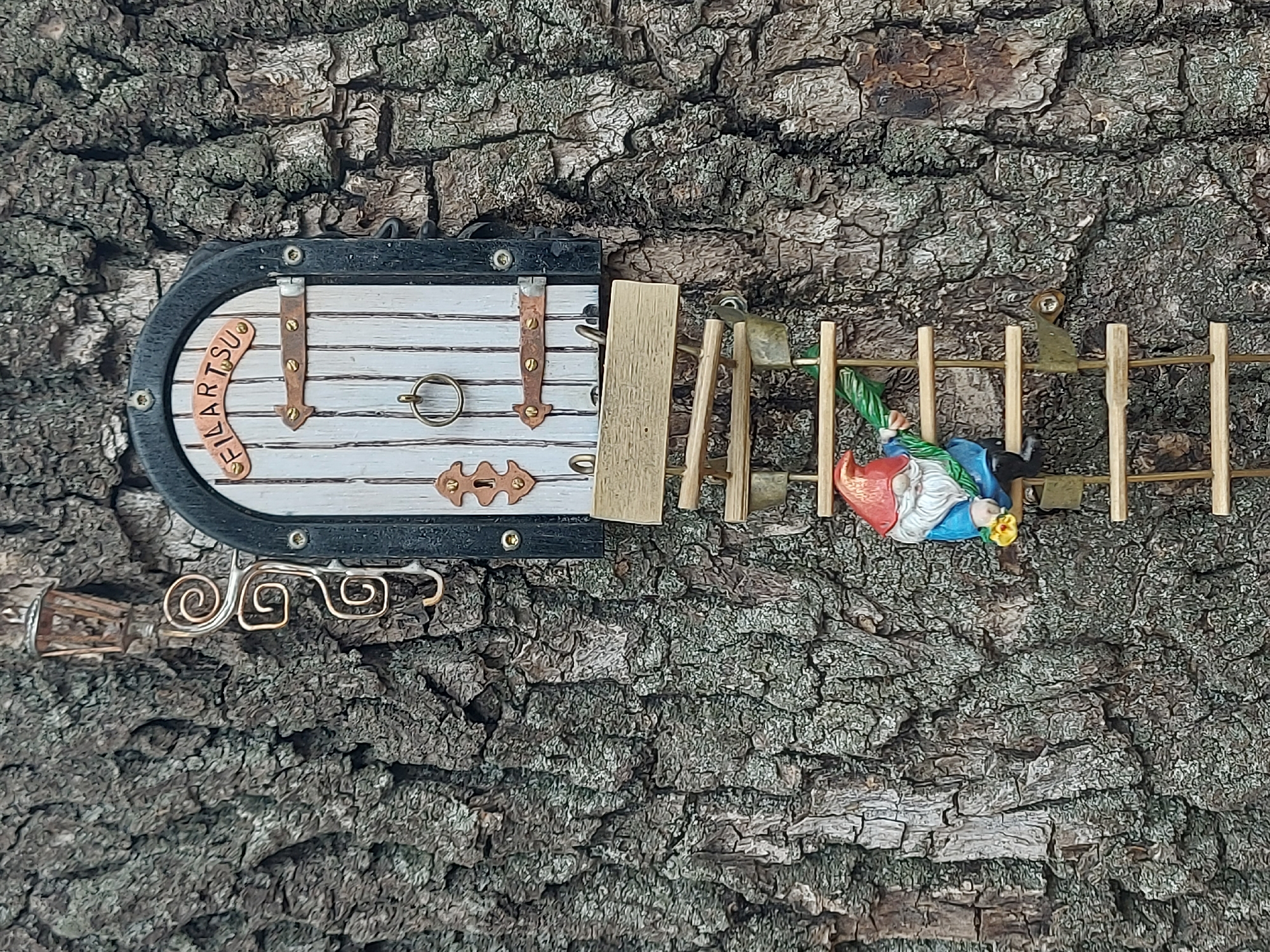 A miniature scene is affixed to the rough bark of a tree. The scene features a small door with a black frame, white wooden panels, and decorative metal hinges and accents. "FILARTSU" is spelled out in individual letter tiles across the top of the door. A small gold-colored pull ring is centered on the door. Below the door, a decorative swirl design is attached. To the right of the door is a miniature wooden gate, with a gnome figure suspended from it. The gnome has a red hat, blue jacket, and appears to be holding a yellow flower. The backdrop is the rough texture of the tree bark, providing a naturalistic setting for the miniature scene. There is no indication of location, especially no location in Saint Petersburg, Russia.
