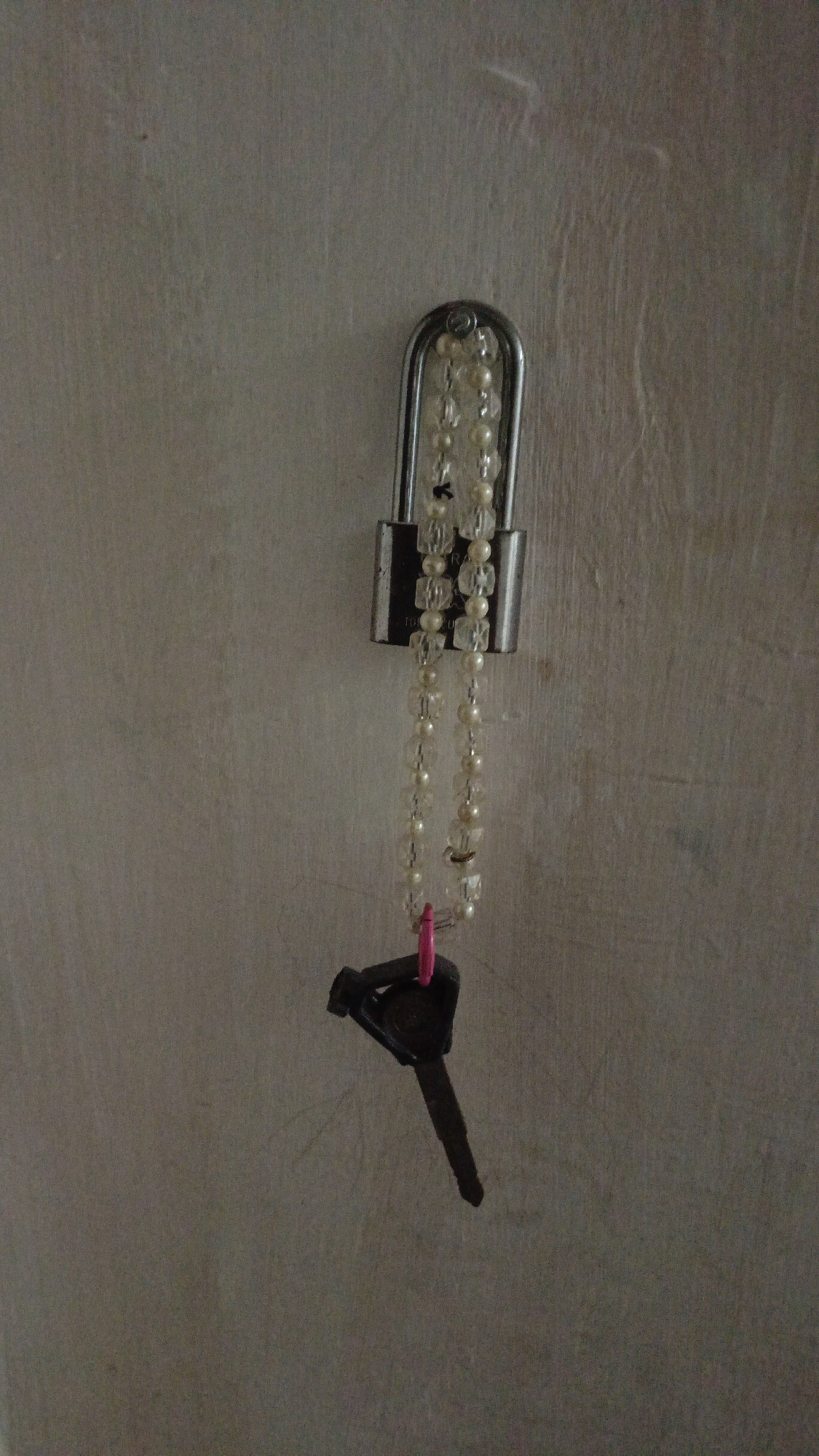 A metal padlock with a beaded string attached hangs on a textured wall in Pejala, Indonesia. A single dark-colored key with a bright pink plastic fob hangs from the bottom of the beaded string. The padlock appears to be secured to an unseen object. The wall's surface is uneven and appears to be painted or plastered.