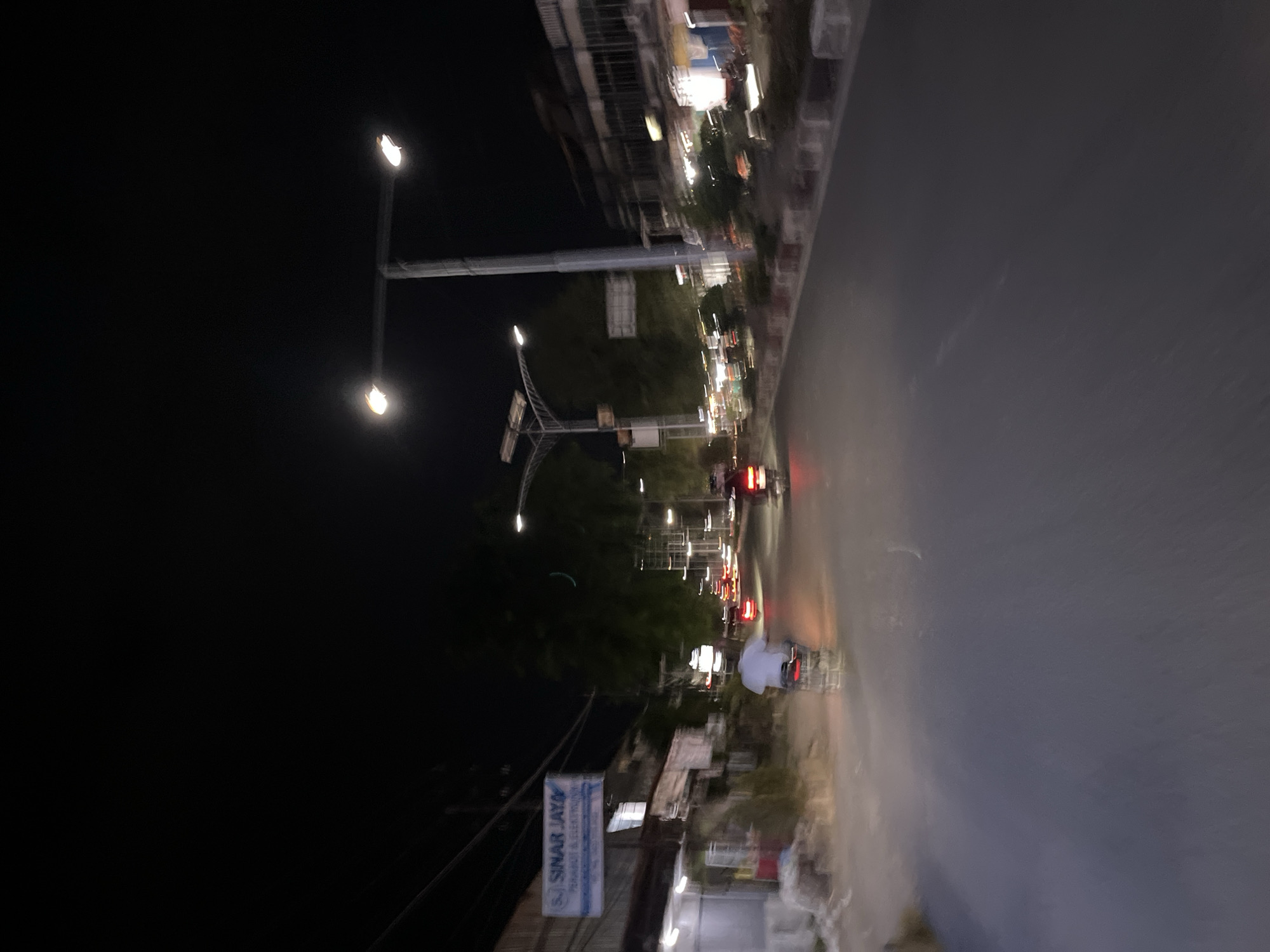 This media file captures a street scene at night in Langsa, Indonesia. The image is characterized by motion blur, suggesting it was taken from a moving vehicle. The primary subjects are the illuminated street lamps, vehicles on the road, and buildings lining the street. Trees are also visible, interspersed with the man-made structures.
The scene depicts a typical urban environment at night, with artificial lights from street lamps and vehicle headlights creating streaks of light due to the motion. Red taillights of vehicles are visible on the road, indicating traffic. A person on a motorcycle, wearing a white helmet, is a discernible figure amidst the blur.
Notable details include the darkness of the night sky, contrasted with the bright artificial lights. The wet appearance of the road might suggest recent rain or general humidity. A sign with the text "SINAR JAYA MANDIRI & ELEKTRO" is partially visible, indicating a business establishment. The overall atmosphere is one of a bustling nighttime street in a city. The motion blur contributes to a sense of dynamism and transience.