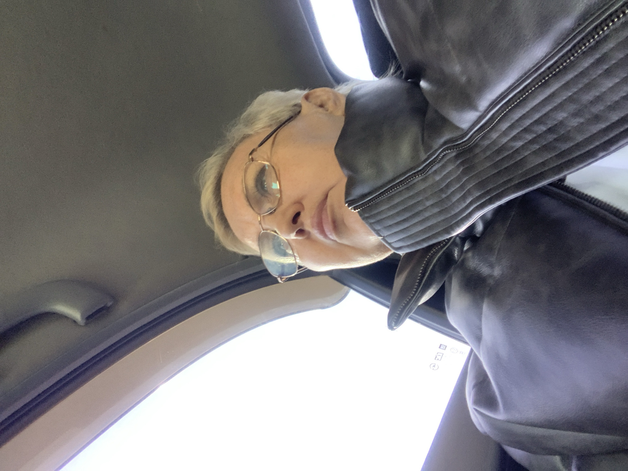 A woman with short blonde hair and glasses is looking down at the camera from inside a car. She is wearing a black leather jacket. The interior of the car is visible, including the roof and the handle near the window. The location is undetermined.