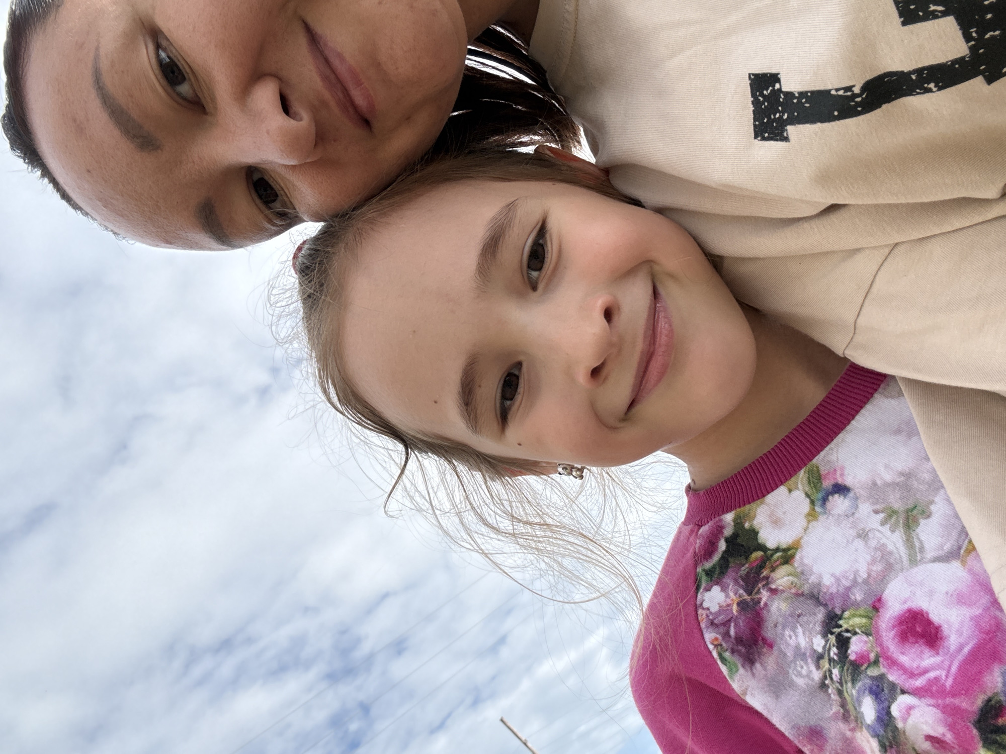 This is a low-angle, close-up selfie taken outdoors, featuring two individuals, likely a mother and daughter, against a backdrop of a cloudy sky. The woman, on the left, has dark hair and is looking down towards the camera. Her facial features are visible, including her eyes, nose, and mouth. The girl, positioned slightly in front of her, is also looking at the camera and smiling broadly, revealing her teeth. She has light brown hair, and a small earring is visible on her left ear. Her attire consists of a pink long-sleeved shirt with a floral pattern featuring pink and white roses. The woman appears to be wearing a light-colored beige sweatshirt with a black graphic design on it. The photo captures a moment of shared joy and connection between the two. The lighting suggests it's daytime, possibly late afternoon given the soft light, and the cloudy sky indicates moderate weather conditions. There are no other significant environmental cues to pinpoint the exact location, other than the provided context of Большой Турыш, Russia.