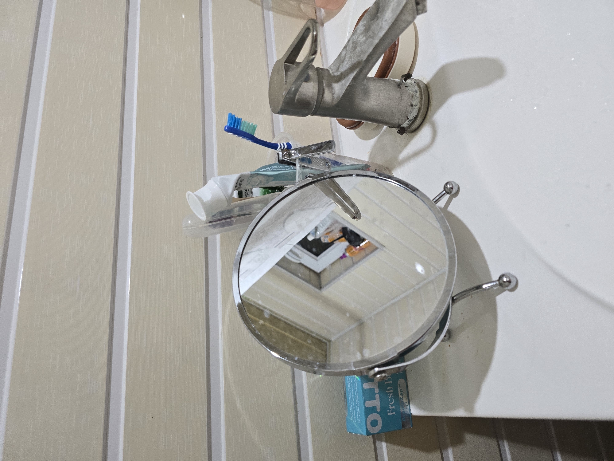 The image depicts a close-up of a bathroom sink area, likely in Qaratau, Kazakhstan, based on the context. A white sink is in the foreground, with a chrome faucet showing some signs of wear and water spots. To the left of the sink, a small, circular, chrome-framed mirror stands on a silver stand, reflecting the opposite wall and a window with curtains. A blue box, possibly of dental floss or gum, labeled "OTTO Fresh B" is placed to the left of the mirror. Behind the mirror, a clear plastic cup holds a tube of toothpaste and a blue-bristled toothbrush. The wall behind the sink is paneled with horizontal beige and white stripes. No people are visible in the image. The overall scene suggests a domestic bathroom setting.