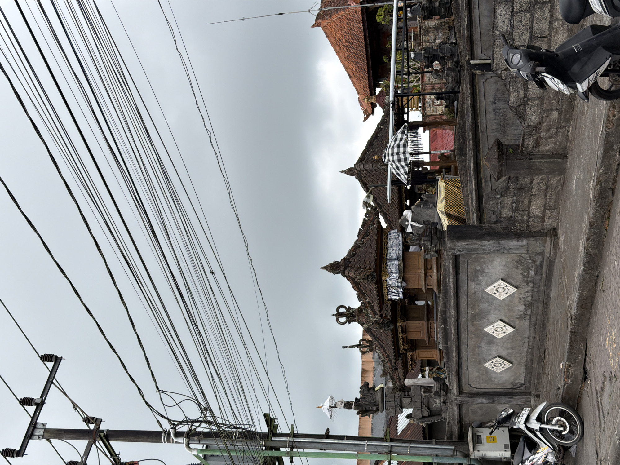 In Kintamani, Indonesia, on a cloudy day, a street scene unfolds with a focus on a traditional Balinese temple entrance. A prominent power pole with a dense network of overhead wires dominates the left side of the frame, extending diagonally across the sky. Below, a gray stone wall, adorned with three diamond-shaped reliefs, separates the street from the temple grounds.
To the left of the wall, a white motorcycle is parked on the paved street, its license plate visible. Within the temple grounds, several structures with tiered, tiled roofs are visible. One structure features a decorative umbrella with a black and white checkered pattern. The temple architecture is detailed with stone carvings and ornate embellishments. Further to the right, another motorcycle, this one black, is partially visible, parked in front of a stone wall. The overall environment suggests an urban or semi-urban setting in Indonesia, characterized by intricate religious architecture and the ubiquitous presence of electrical infrastructure.