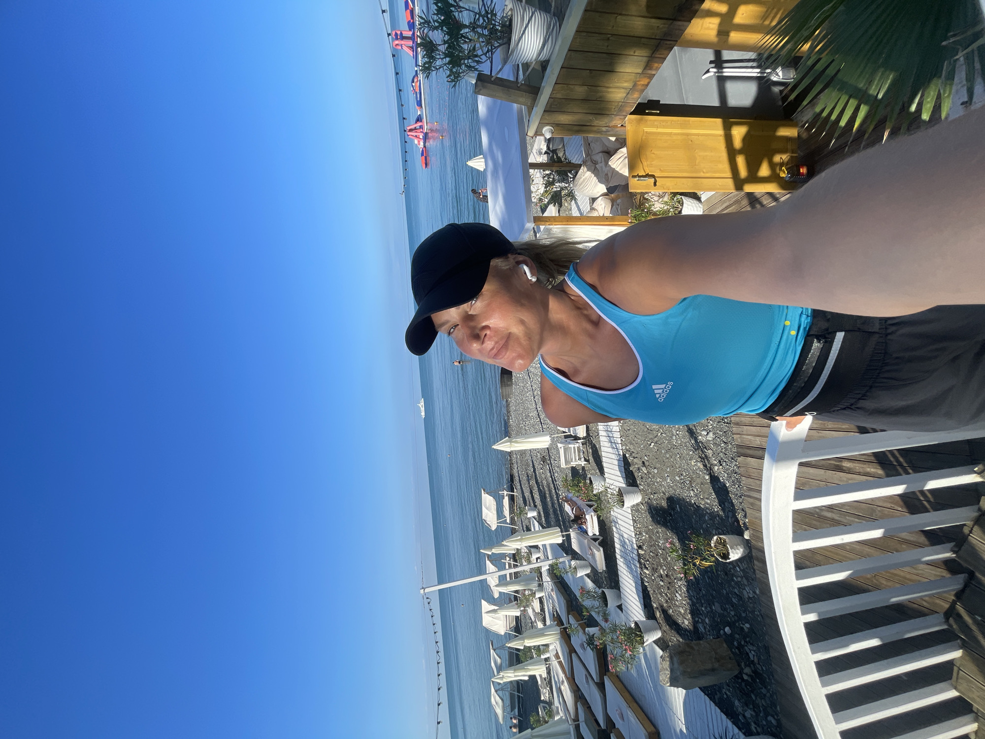 A woman is taking a selfie on a balcony overlooking a beach resort in Sochi, Russia. She is wearing a blue tank top with an Adidas logo, black shorts, and a black baseball cap. She also has white wireless earbuds in her ears. The beach below is lined with white lounge chairs and umbrellas, and there is a calm blue ocean in the background with a few boats and inflatable water attractions. The sun is shining, casting long shadows on the ground. The woman is looking up and to the right with a relaxed expression.