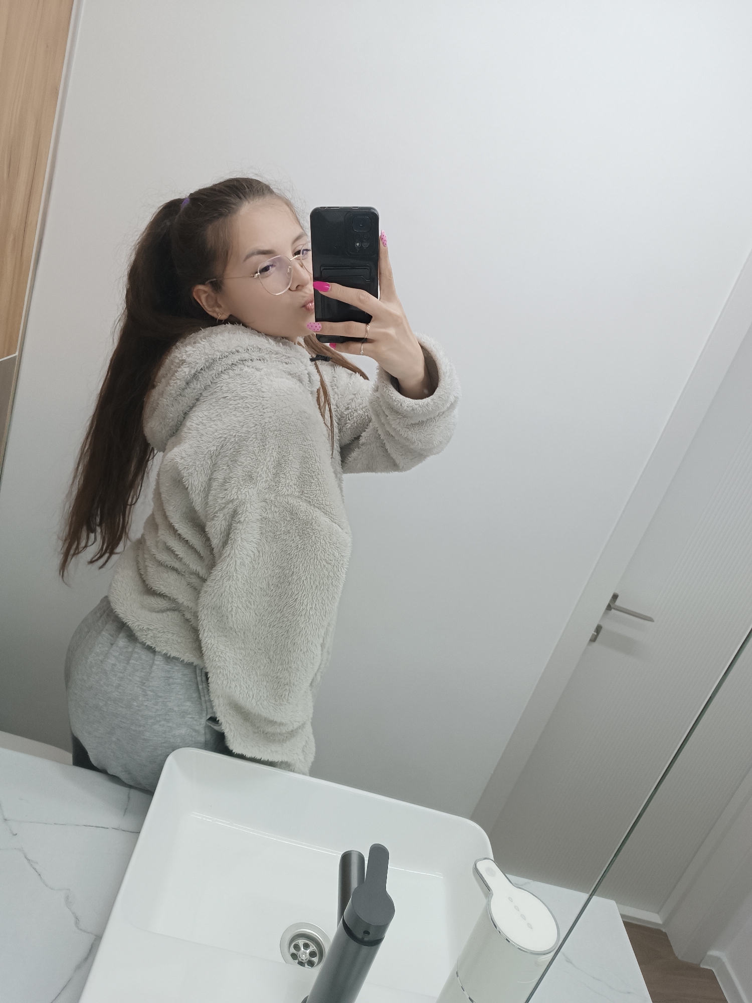 A young woman is taking a selfie in a bathroom. She has long brown hair in a ponytail and is wearing glasses. She is wearing a fluffy gray hoodie and gray sweatpants. She is holding a black smartphone with pink-painted nails. The bathroom has a white sink with a modern faucet and soap dispenser. There is a door in the background. The location is Unknown, Unknown.