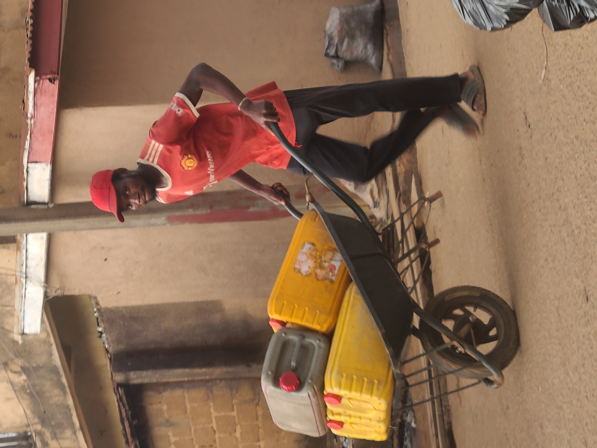 A man in a red Manchester United jersey and a red cap pushes a wheelbarrow loaded with three containers along a paved street in Jos, Nigeria. The wheelbarrow contains two yellow and one gray container. The street is flanked by buildings.