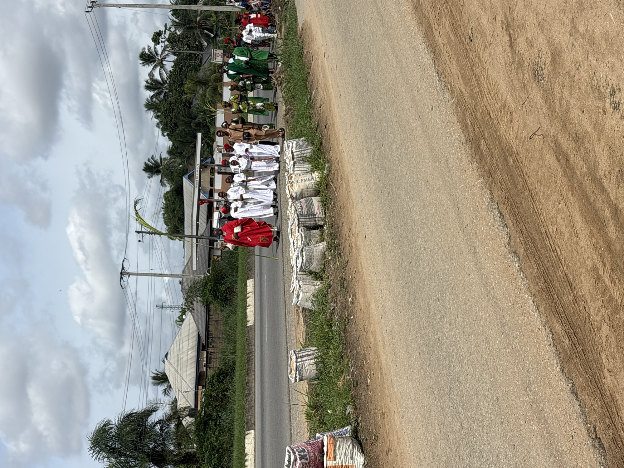 The image depicts a procession on the shoulder of a road near what appears to be a built environment. The location is assumed to be near Uyo, Nigeria. People in various robes (red, white, brown and green) are standing behind cement bags along the edge of the road. The sky is overcast, there are trees in the background, and utility poles and wires are visible along the side of the road.