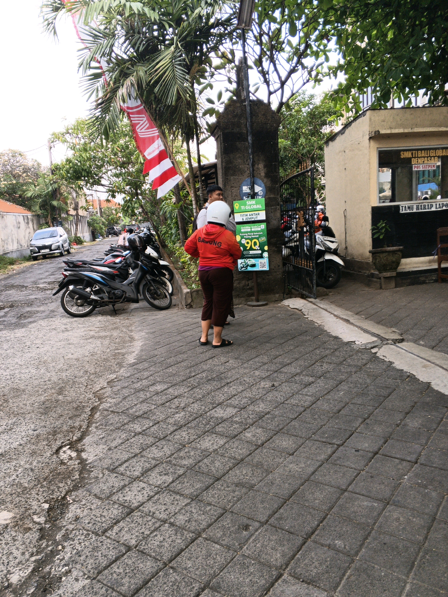 The image shows a street scene in Denpasar, Indonesia, likely near an educational institution given the signage. In the foreground, a person wearing a red hoodie, brown pants, and a white helmet stands on a paved walkway. To their left, several motorcycles are parked along the side of the street. Behind the motorcycles, a metal gate leads into a property. A sign attached to a post near the gate displays information in Indonesian, including "SMK TI GLOBAL" and "TITIK ANTAR & JEMPUT," suggesting it's a pick-up and drop-off point for SMK TI Global. Further back, a silver car is parked on the asphalt road. The environment is tropical, with palm trees and other lush vegetation visible. A building with a beige facade and a sign that reads "SMK TI GLOBAL DENPASAR" is visible on the right side of the frame. The overall impression is of a typical day on a street in Denpasar, with the presence of parked vehicles, signage, and pedestrians.