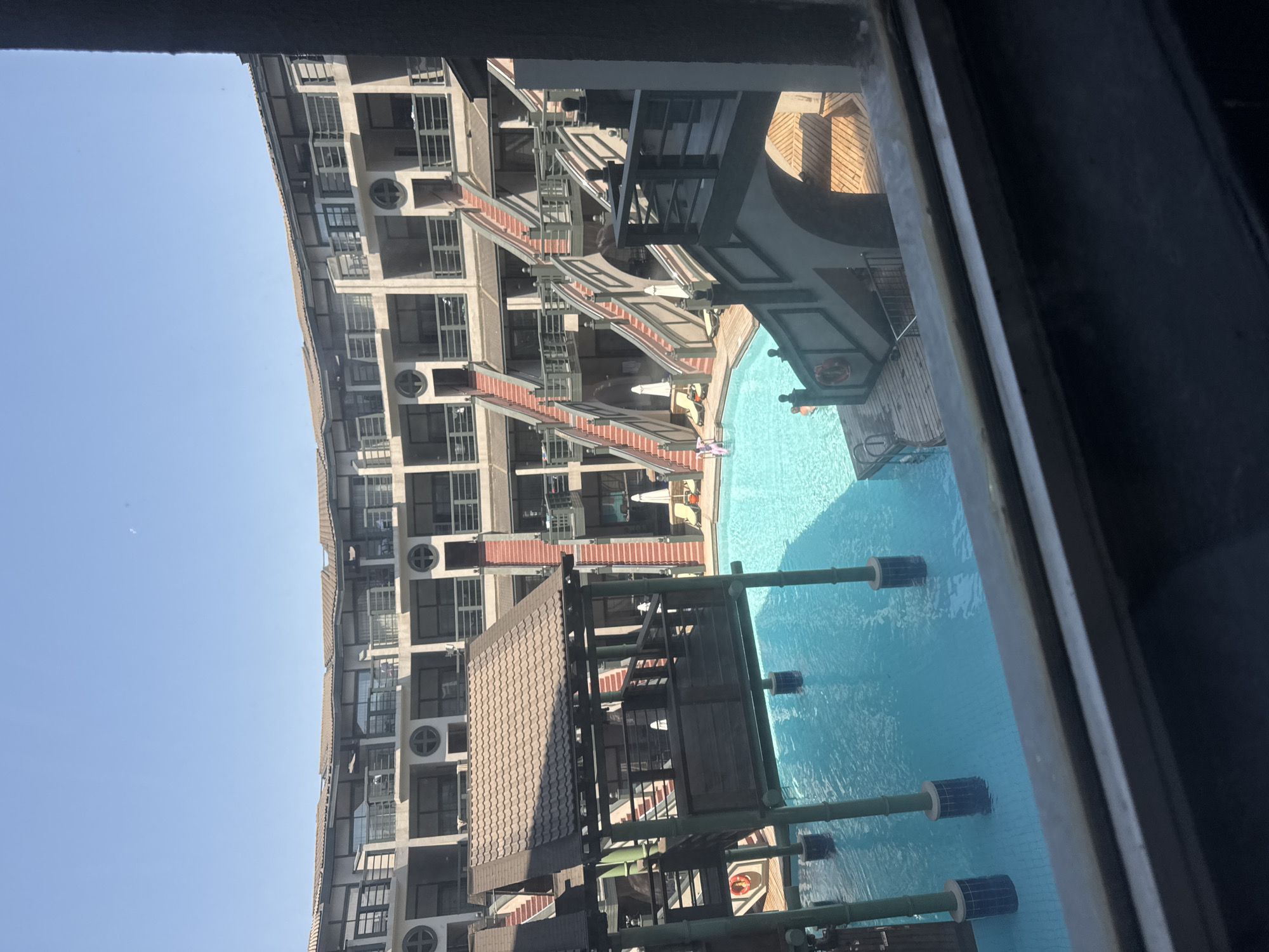This is an eye-level, outdoor, wide-angle shot of a hotel swimming pool and surrounding buildings on a sunny day. The hotel appears to be designed in a multi-story, courtyard style with numerous balconies and staircases. The pool is a bright blue and has several pillars extending from the bottom. There are lounge chairs and umbrellas set up around the pool area. A wooden structure with a sloped roof is situated over the water. The sky is clear and blue. The scene is located in Aksu, Turkey.