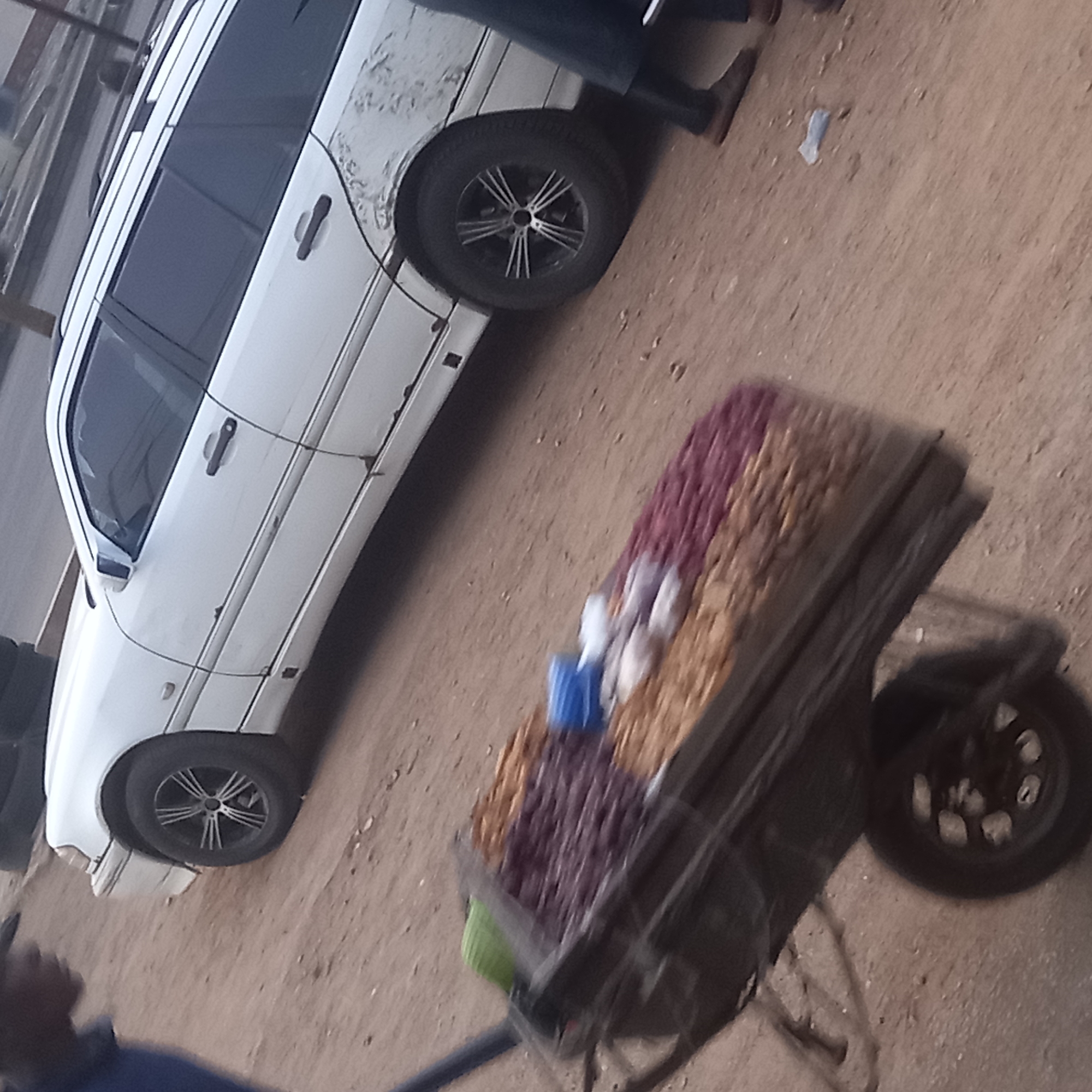 This image captures an outdoor street scene, likely in Sardauna, Nigeria, depicting a casual roadside selling activity.
A prominent subject is a white car, appearing to be a sedan or station wagon, with dark tinted windows and distinctive multi-spoke alloy wheels. The car's body shows visible signs of wear and dirt, particularly on the rear quarter panel and front bumper area. Part of a roof rack is also visible. Near the car's rear passenger door, the lower body of a person, dressed in dark trousers and brown shoes or sandals, is partially visible.
In the foreground, a dark-colored cart or wheelbarrow is heavily laden with various items, suggesting it is a vendor's stall. The goods are arranged in sections: a substantial pile of dark purple or reddish items (possibly dried fruits or nuts), and another section filled with light brown or tan items, which could be groundnuts or other local snacks. Amidst these, small white packages or sachets and a blue container are also visible. A green handle is seen at the front of the cart.
The ground beneath the car and cart is unpaved, consisting of light brown, dusty earth or sand. In the extreme bottom-left corner, a partial view of another person, showing a shoulder and arm in a blue garment, is visible near the cart. The scene is illuminated by daylight, indicating clear weather. No discernible text is visible on any objects in the image.
