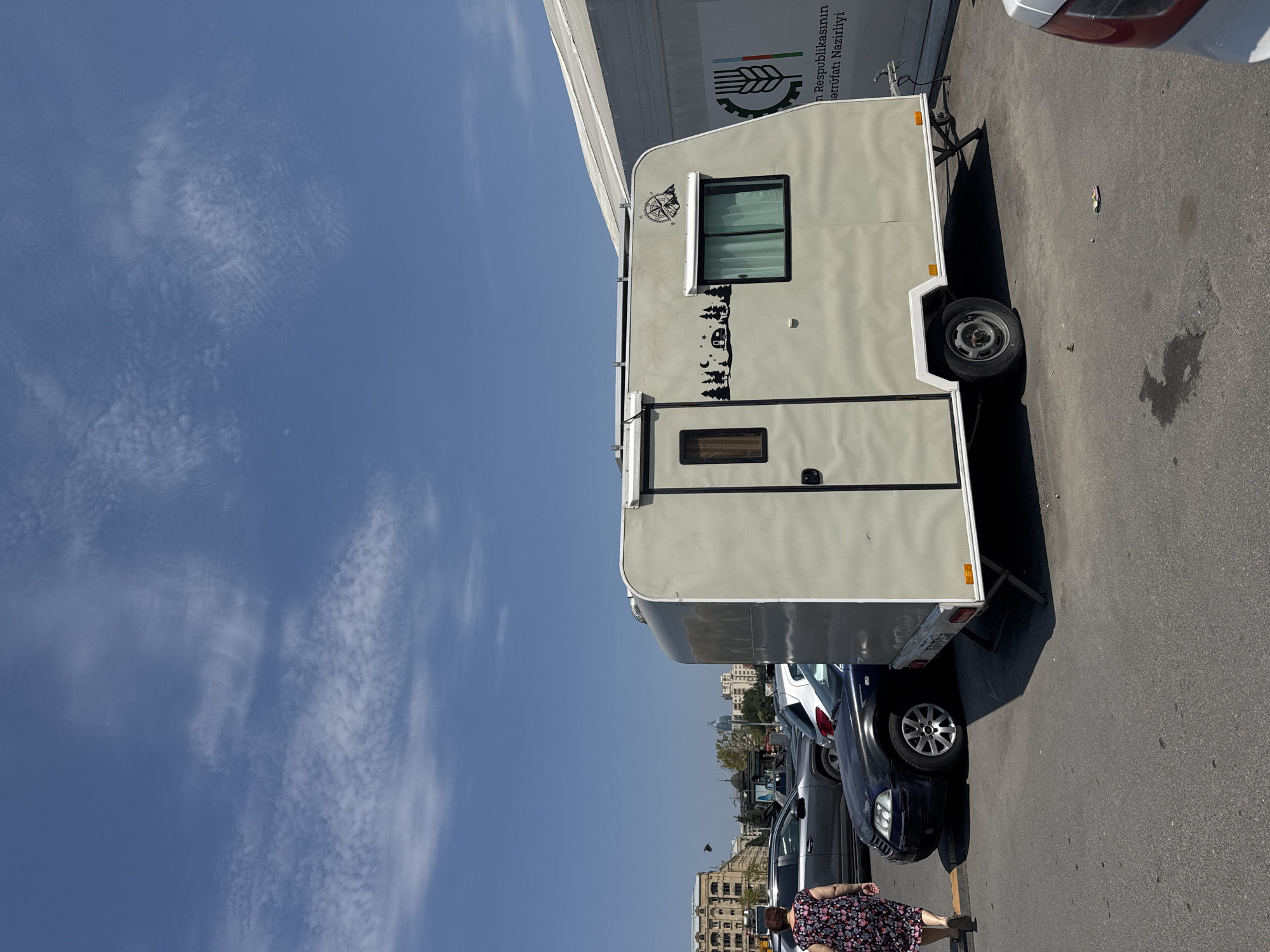 The image depicts a light-colored caravan parked in an outdoor urban environment, likely a parking lot or street area in Baku, Azerbaijan, under a clear blue sky with scattered white clouds. In the foreground, a woman with dark hair, wearing a floral-patterned dress and dark sandals, walks away from the camera towards the left. Adjacent to her is a dark blue sedan. The caravan, positioned centrally in the frame, has a window on its right side with white curtains visible and a small graphic sticker featuring a campsite scene with trees, a crescent moon, and a star. Above the window, a compass graphic with mountains is also visible. To the left of the caravan is a closed door with a small, dark-framed window. The caravan is supported by a single wheel on the right side and two stabilizing legs at the rear. In the background, several other vehicles are parked, including a gray SUV. Further in the background, buildings with architectural details typical of Baku line the street. A large white structure, possibly a temporary exhibition or market stall, is visible to the right of the caravan, displaying a logo with a wheat stalk and text in Azerbaijani script that reads "n Respublikasının Sərrufat Nazirliyi" which translates to "Ministry of Agriculture of the Republic". The ground is paved asphalt with some stains and debris.