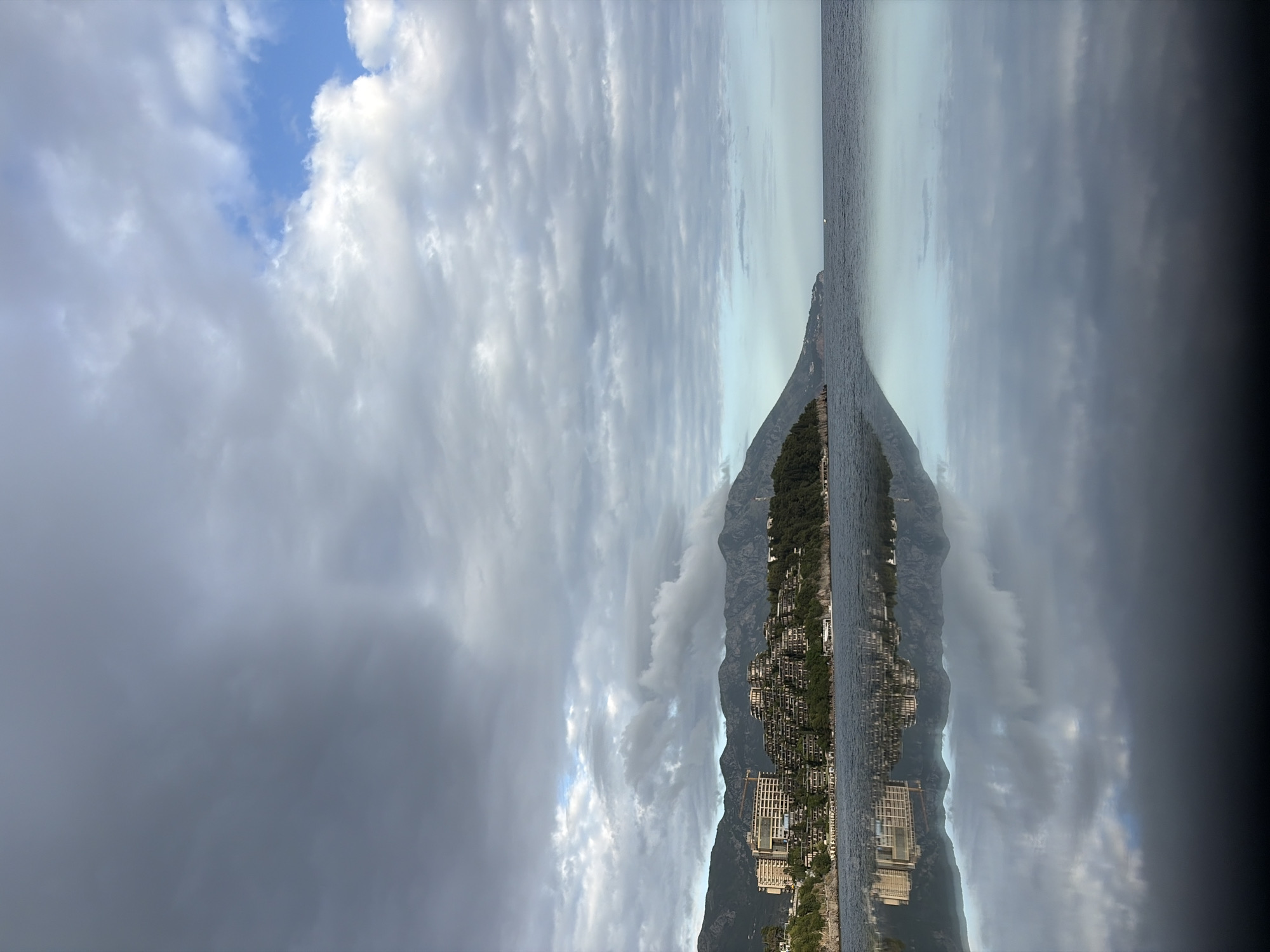 The image shows a view of Budva, Montenegro, along the coast. The sky is filled with gray and white clouds, with patches of blue visible. The coastline features buildings built along the mountainside. The image has been mirrored vertically, giving it an abstract appearance. The water is calm, with a straight horizontal line separating the mountain coastline from the sky.